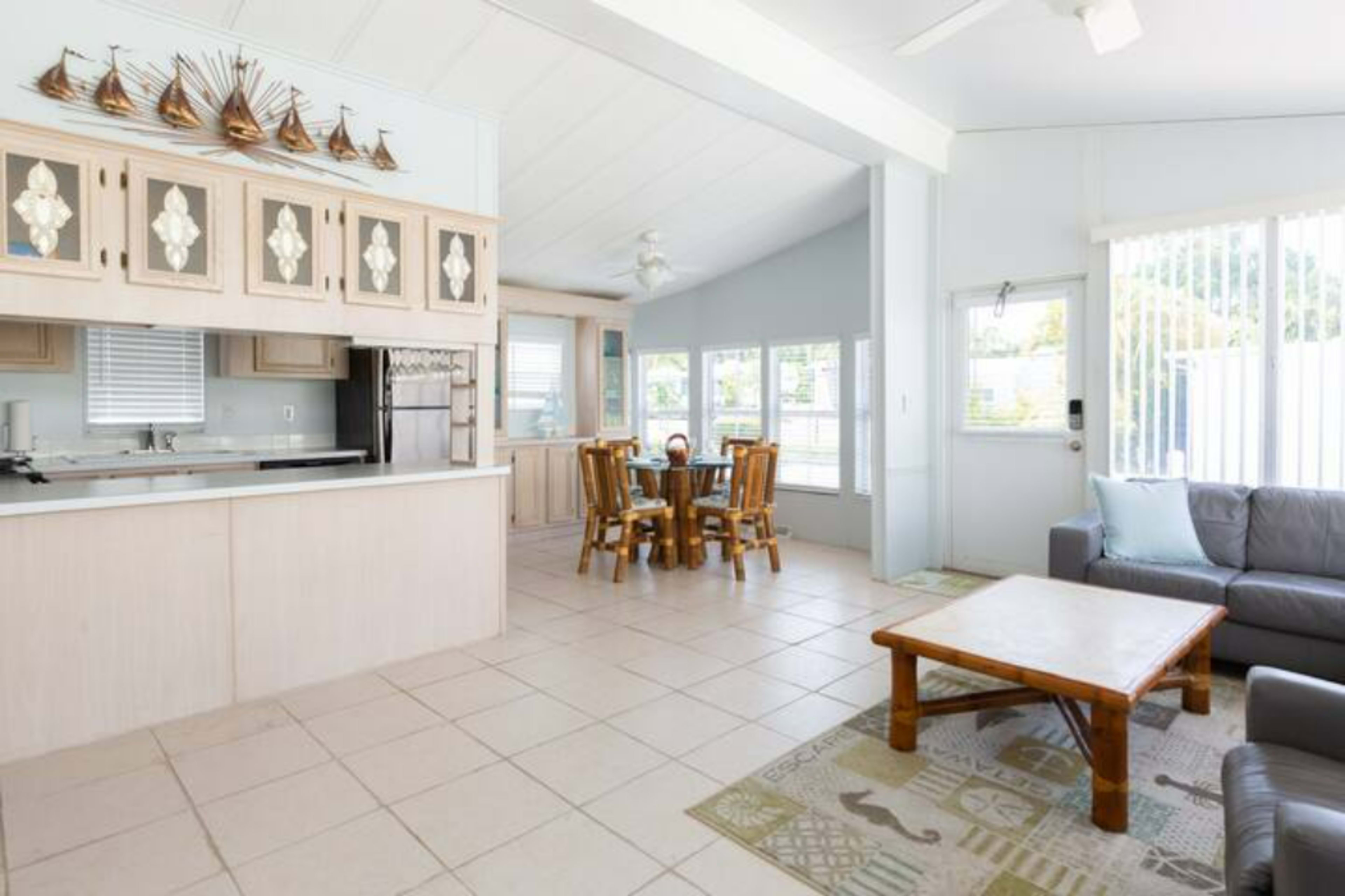 A living area features a sofa and coffee table, leading to a dining space with a round table and chairs, all within a brightly lit room with large windows.