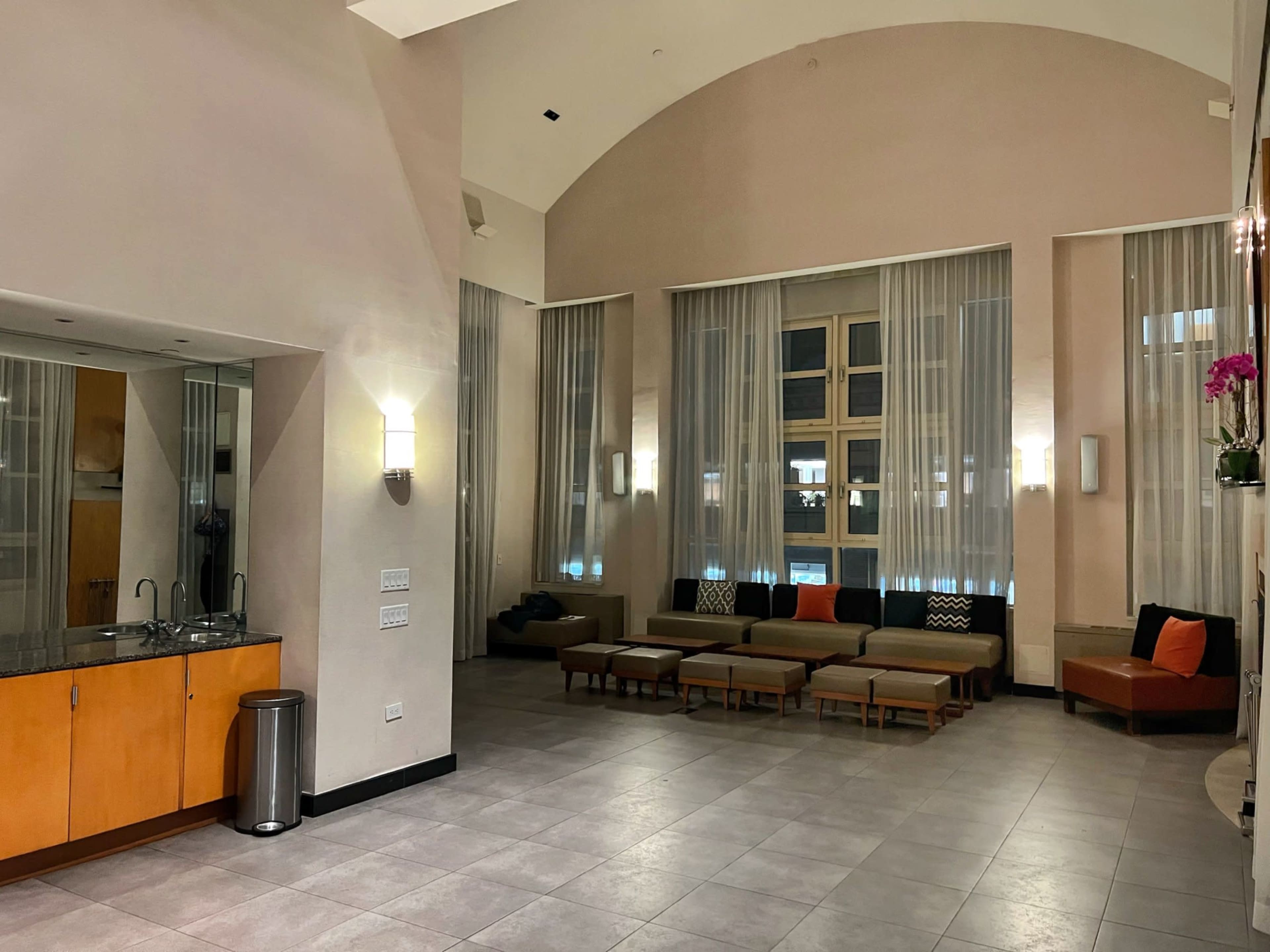 The image shows a modern lounge area featuring a seating arrangement with cushions, large windows with sheer curtains, and a kitchenette with a countertop.