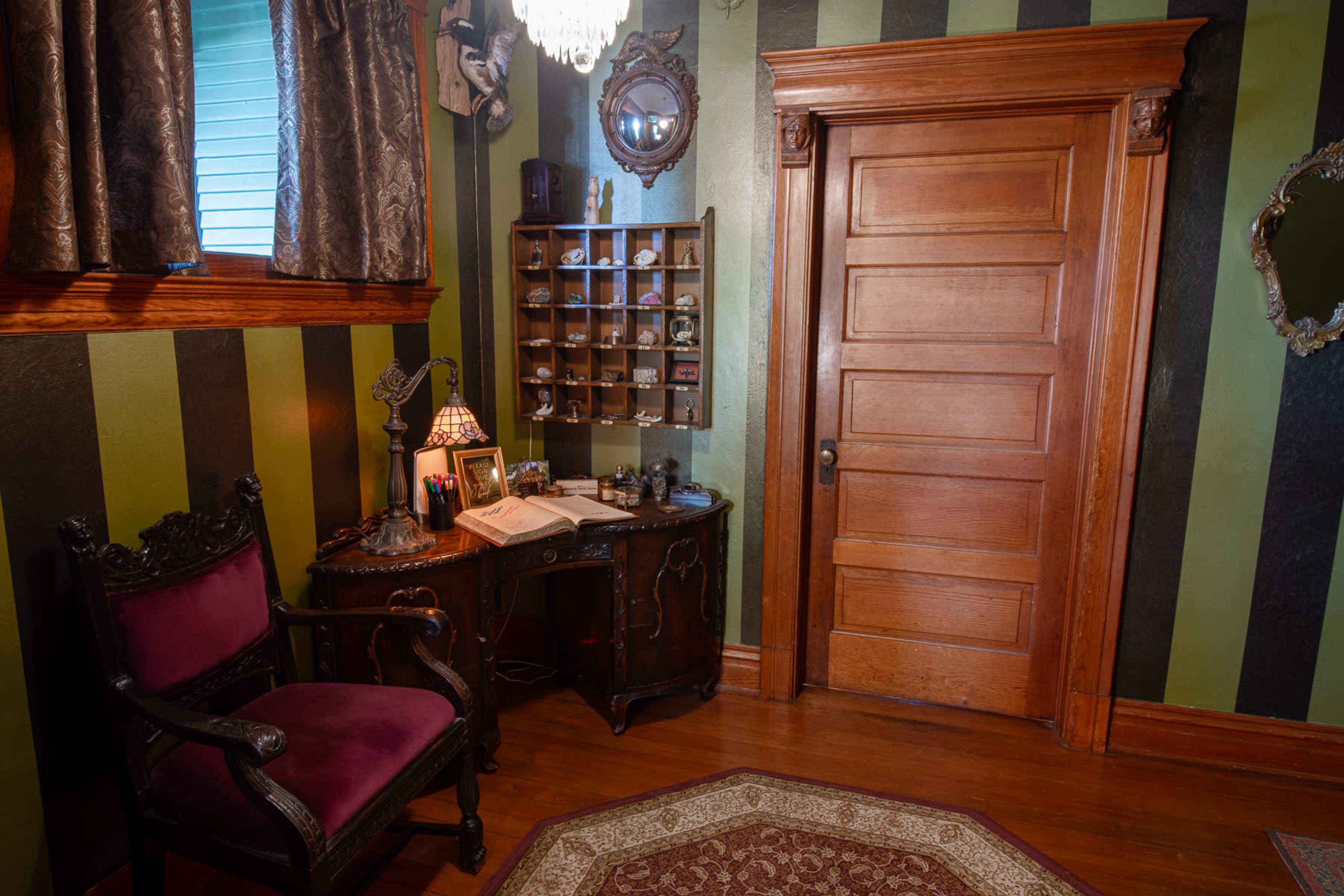 A corner of a room features a dark wooden desk with a lamp and a book, alongside a chair, against a wall decorated with vertical green and black stripes and a wooden door.