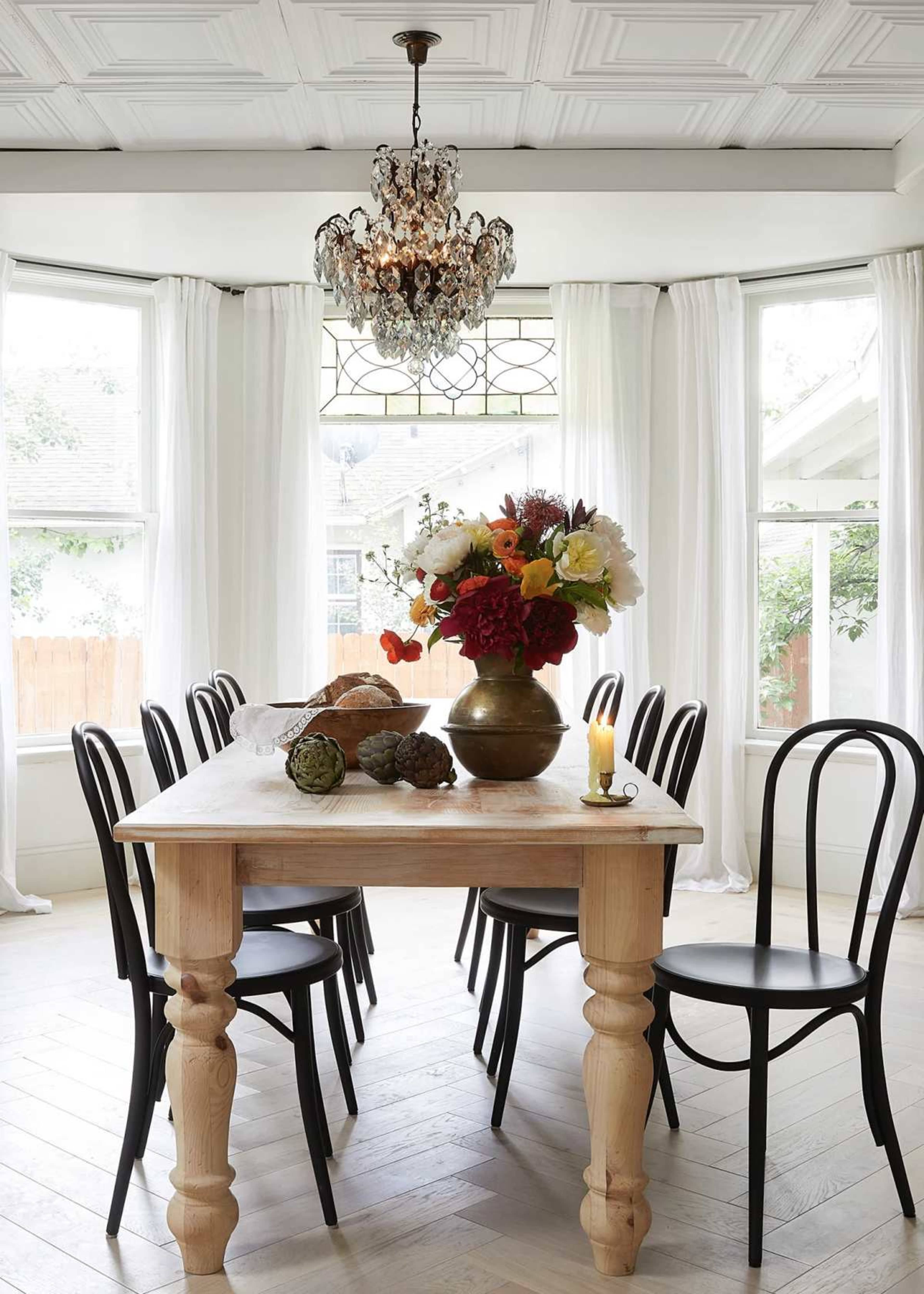 A wooden dining table is set in a bright room with large windows, surrounded by black chairs and adorned with a vase of flowers and various decorative items.