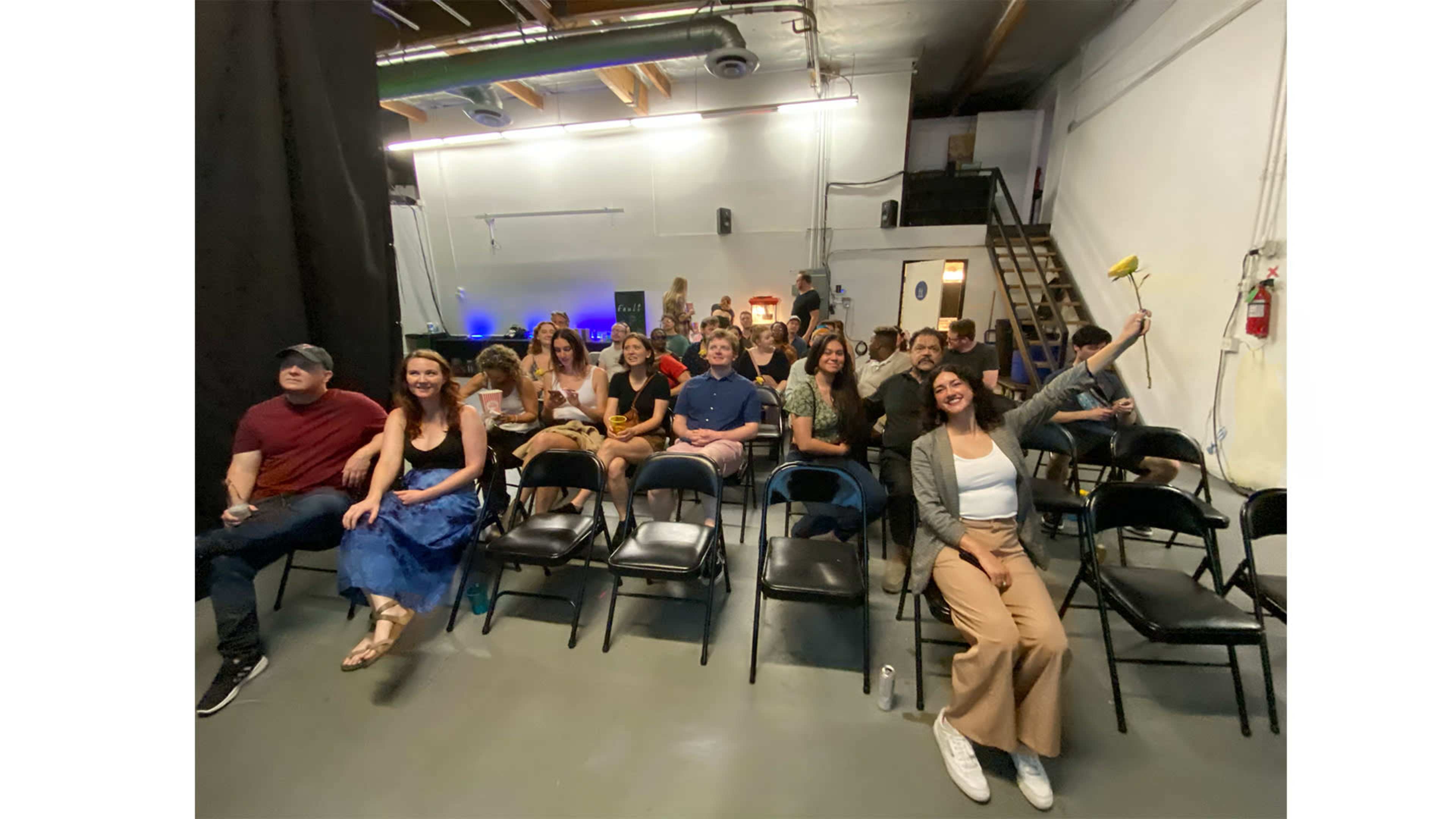A group of people sits in rows of black chairs in a dimly lit space, with a staircase visible in the background.
