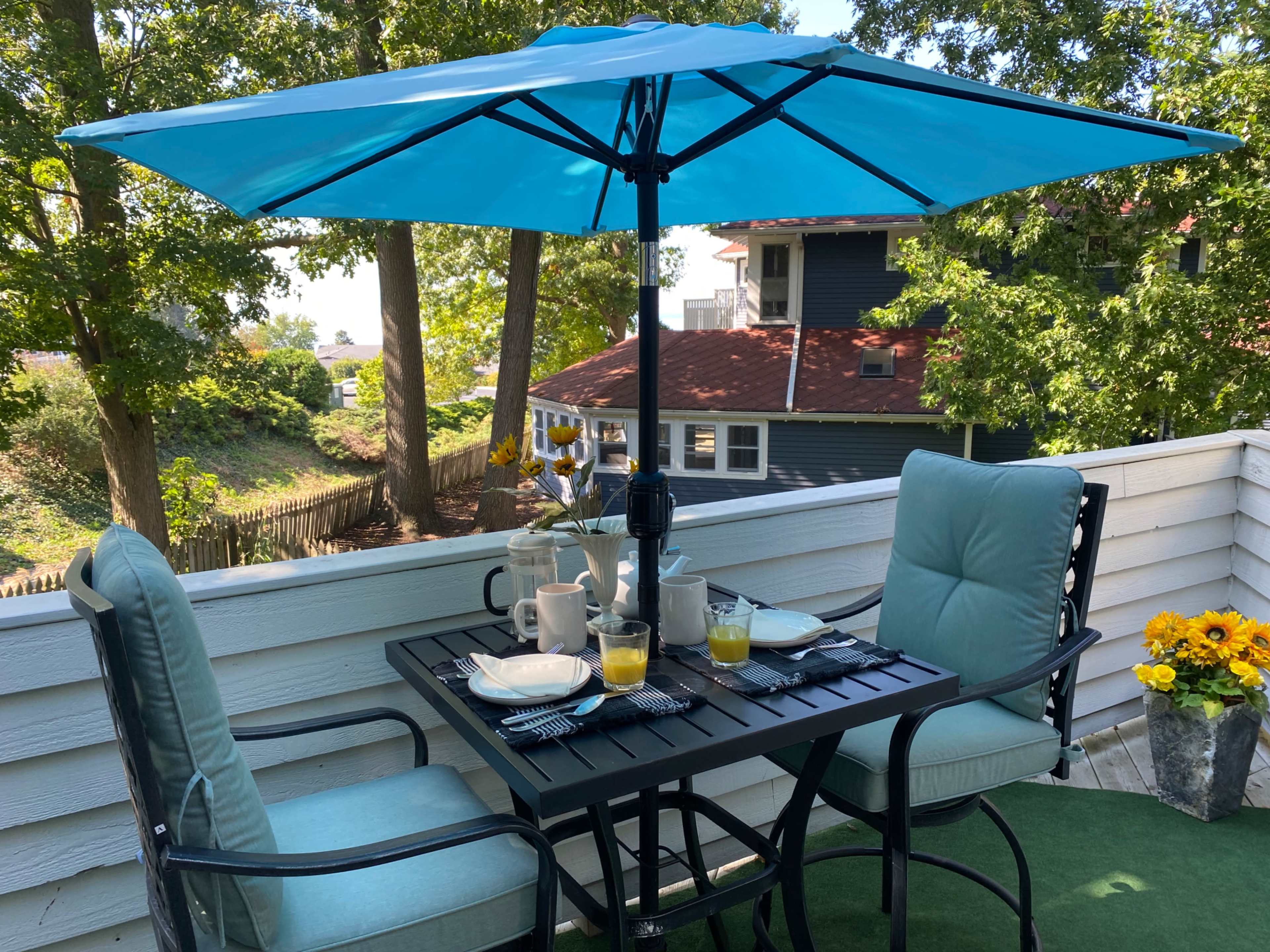 A table set for breakfast is positioned under a turquoise umbrella on a balcony surrounded by trees.