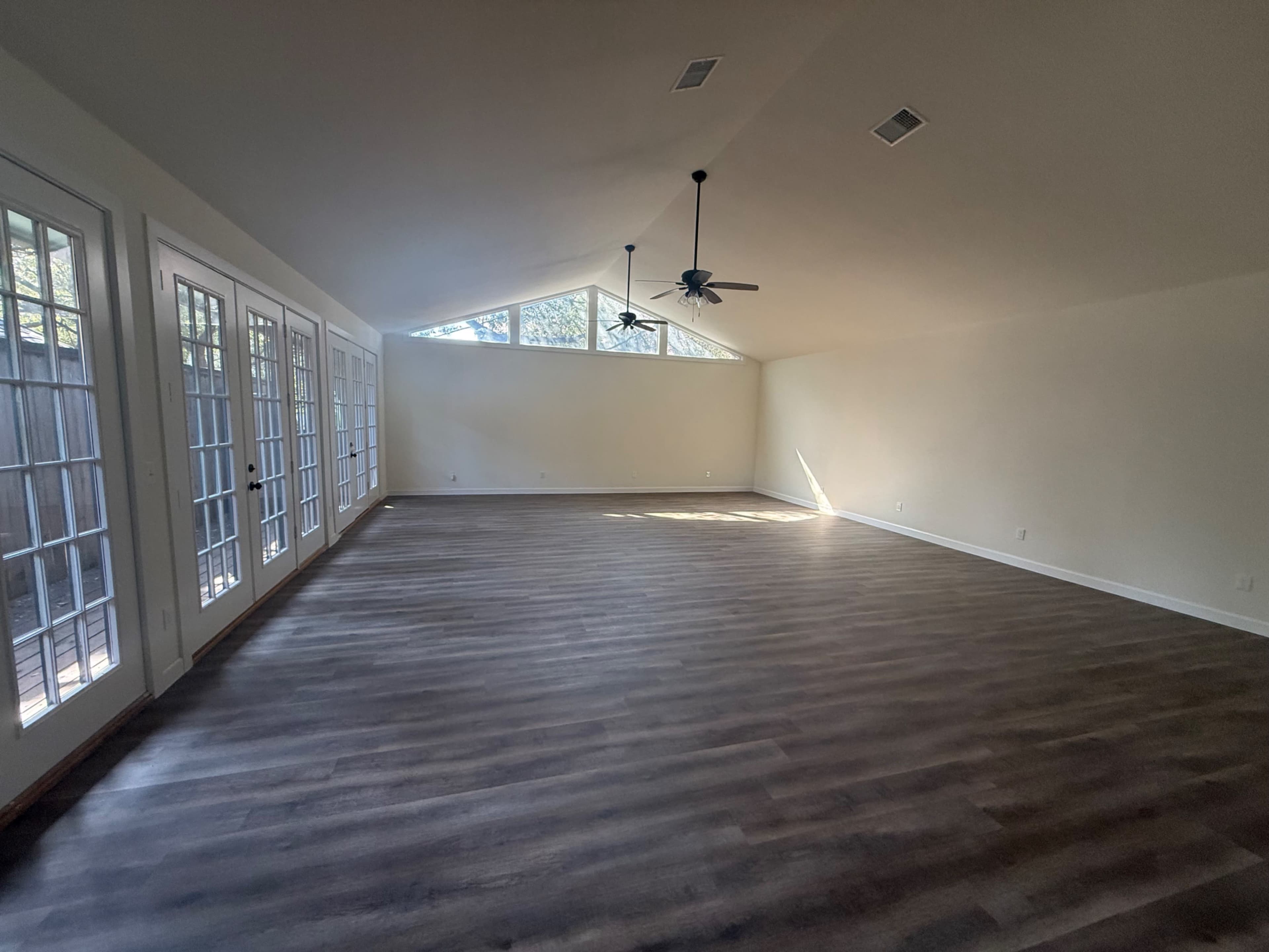 The image shows a spacious room with a high ceiling, featuring large windows and dual ceiling fans.