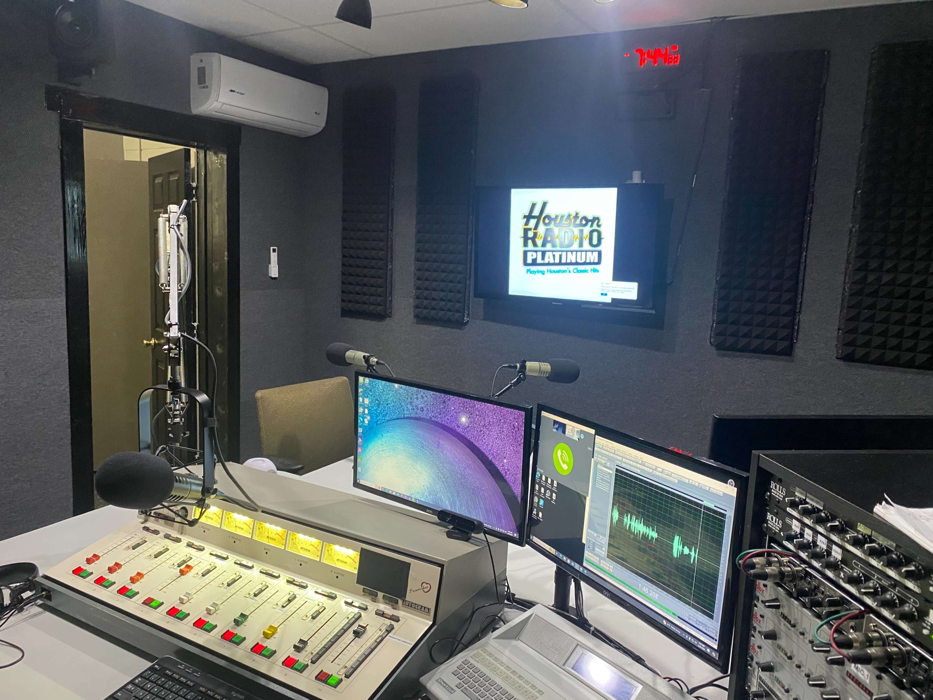 A radio studio equipped with microphones, two computer monitors, a sound mixing board, and soundproofing panels on the walls.