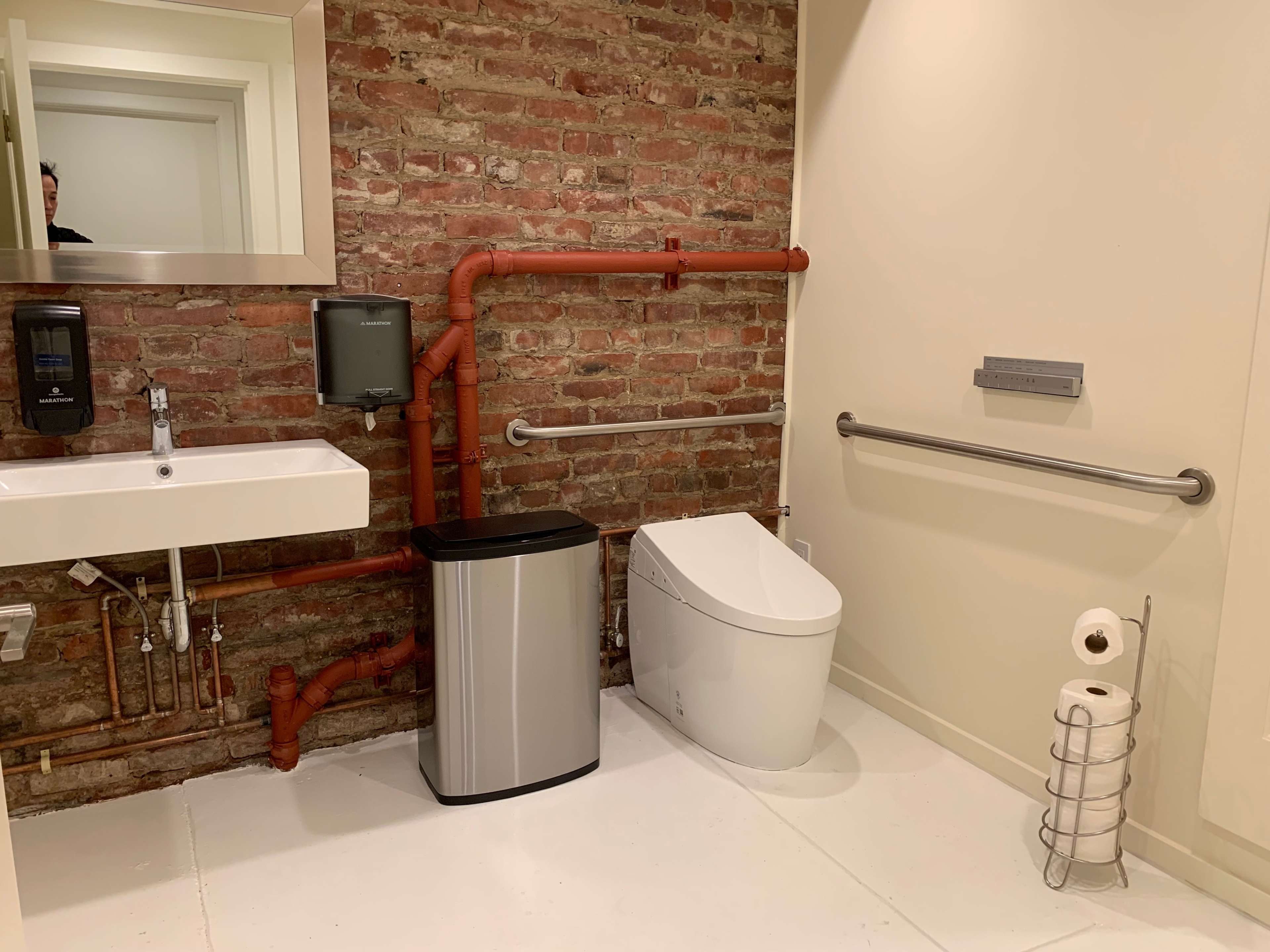 The image shows a bathroom featuring exposed brick walls, a wall-mounted sink, a modern toilet, a trash can, and plumbing pipes along the wall.