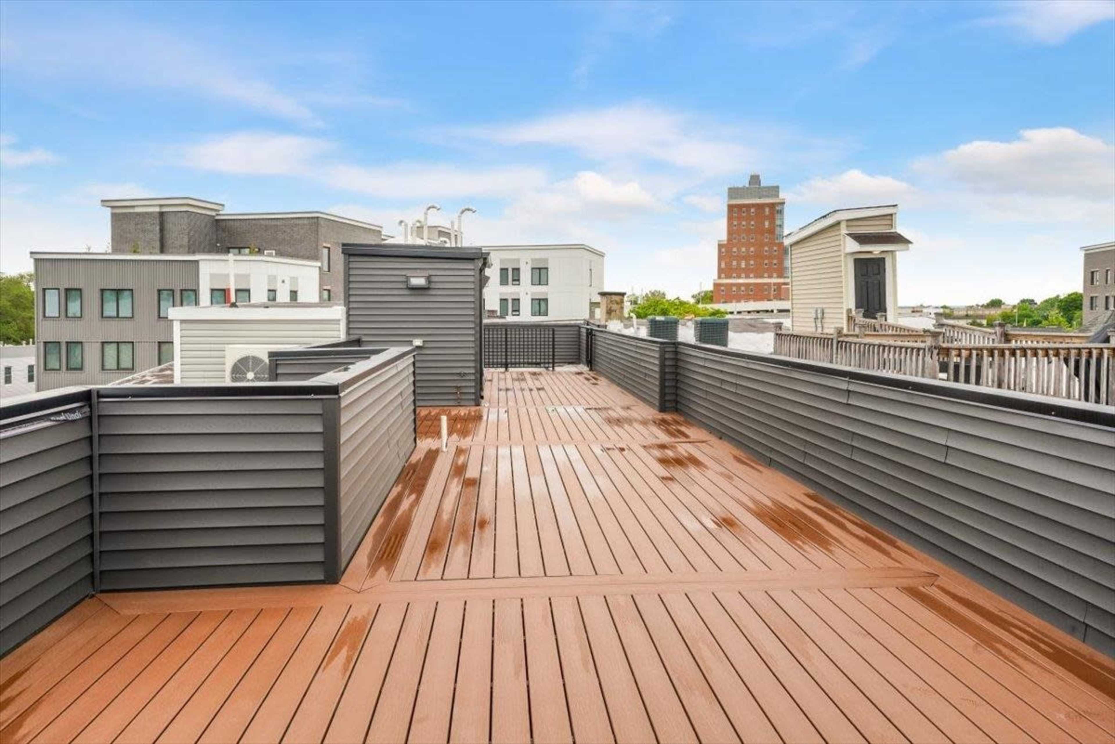 The image shows a spacious rooftop deck with wooden flooring and modern buildings in the background under a clear sky.