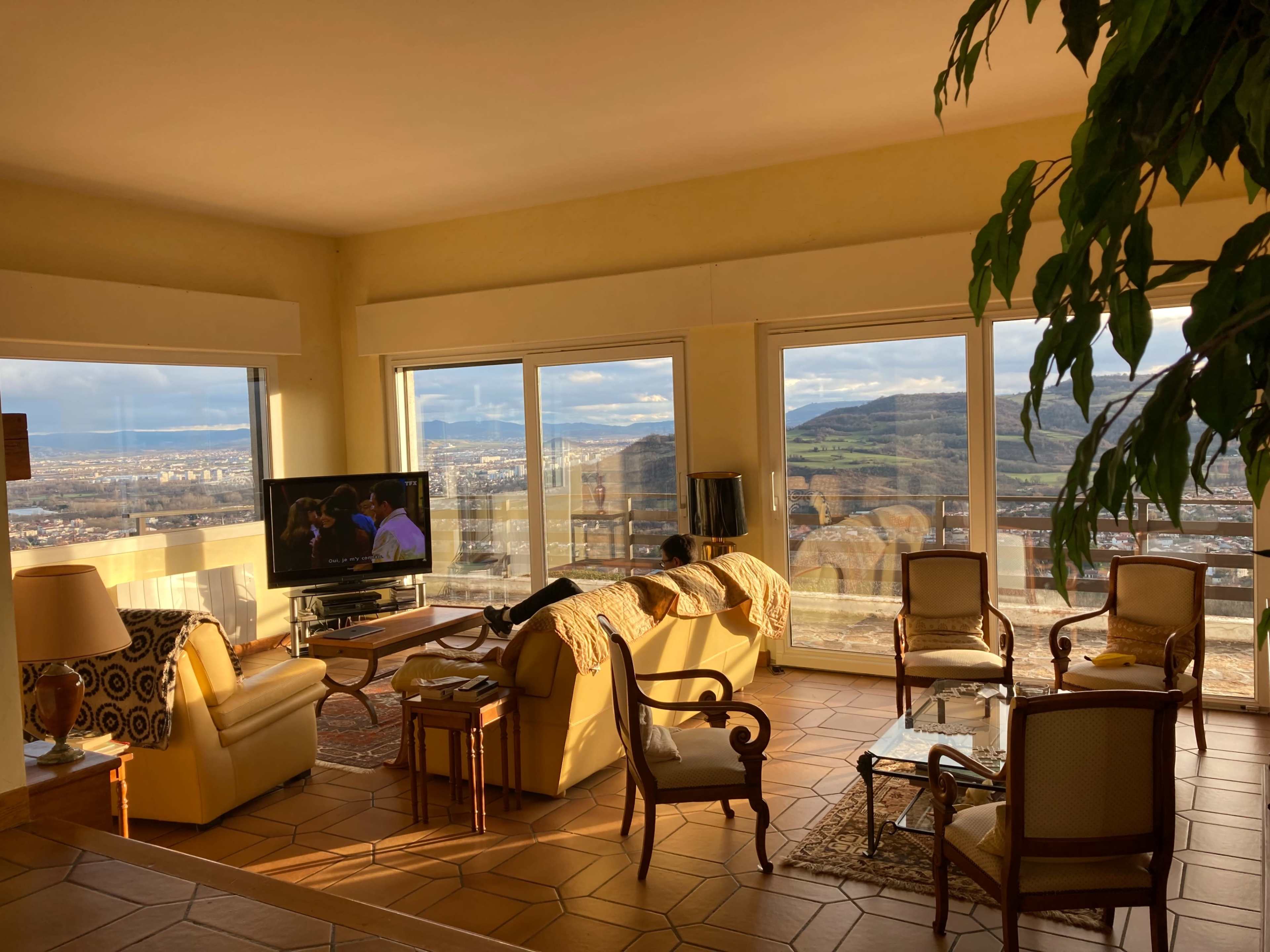 The image shows a living room with large windows overlooking a cityscape, featuring beige furniture, a television, and a plant.