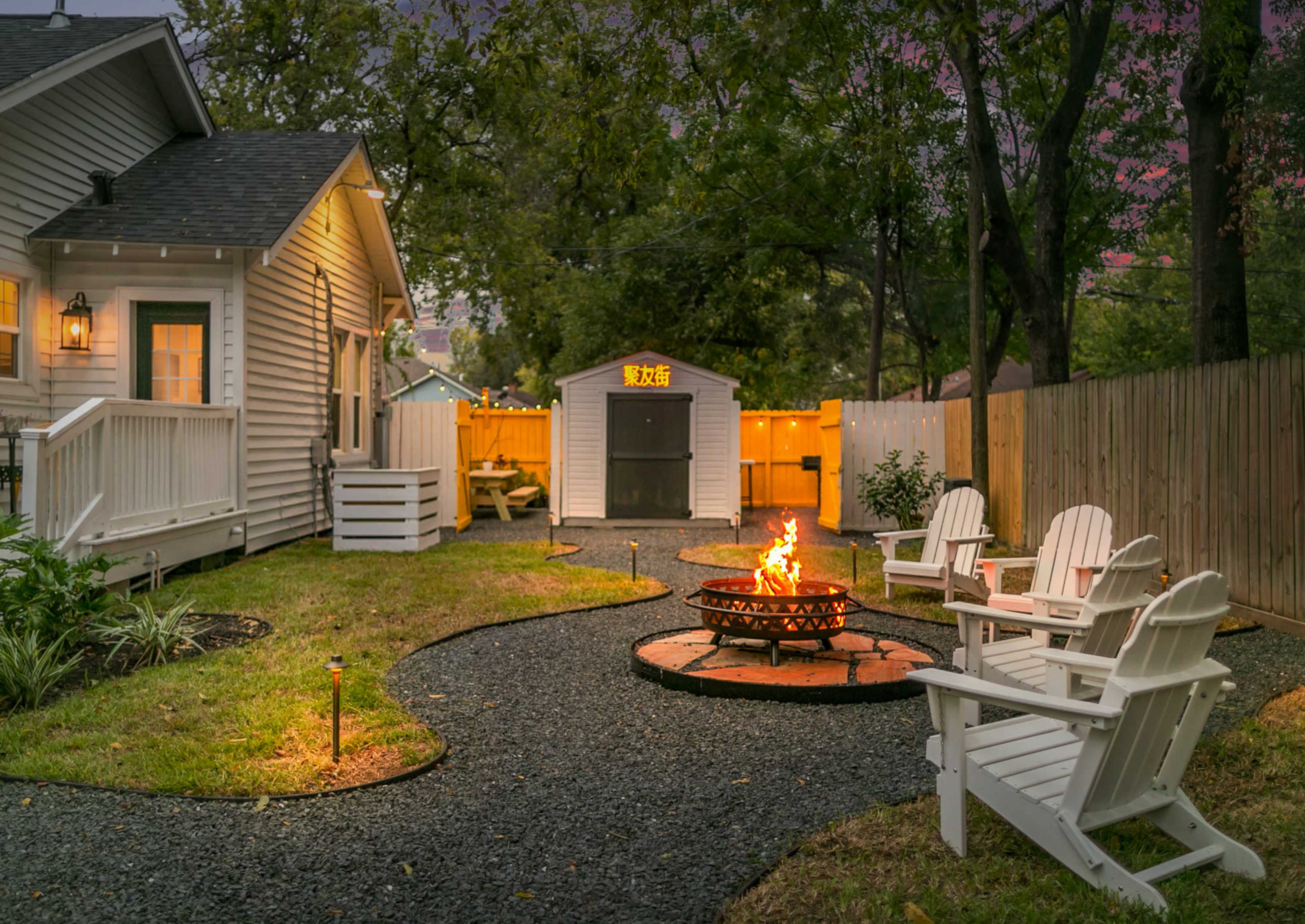 A cozy backyard features a fire pit surrounded by four white chairs, with a small shed visible in the background and lush greenery nearby.