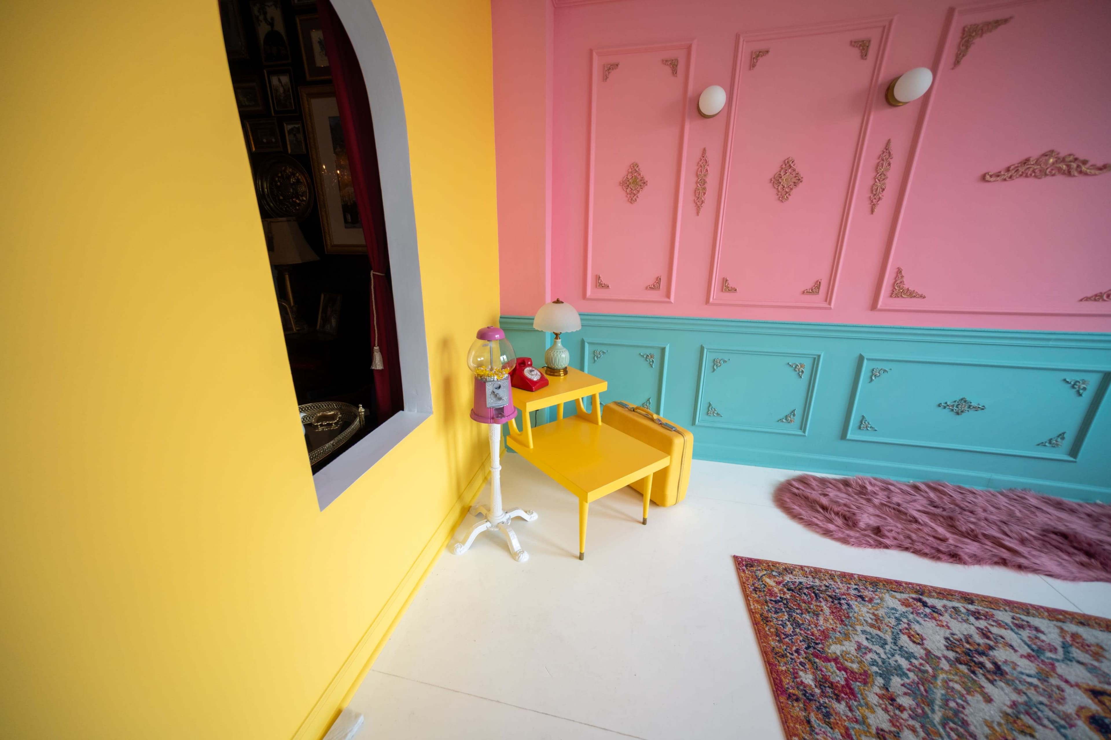 The image shows a brightly colored room featuring yellow and pink walls, with a turquoise panel, a small yellow table, a lamp, and a decorative rug.