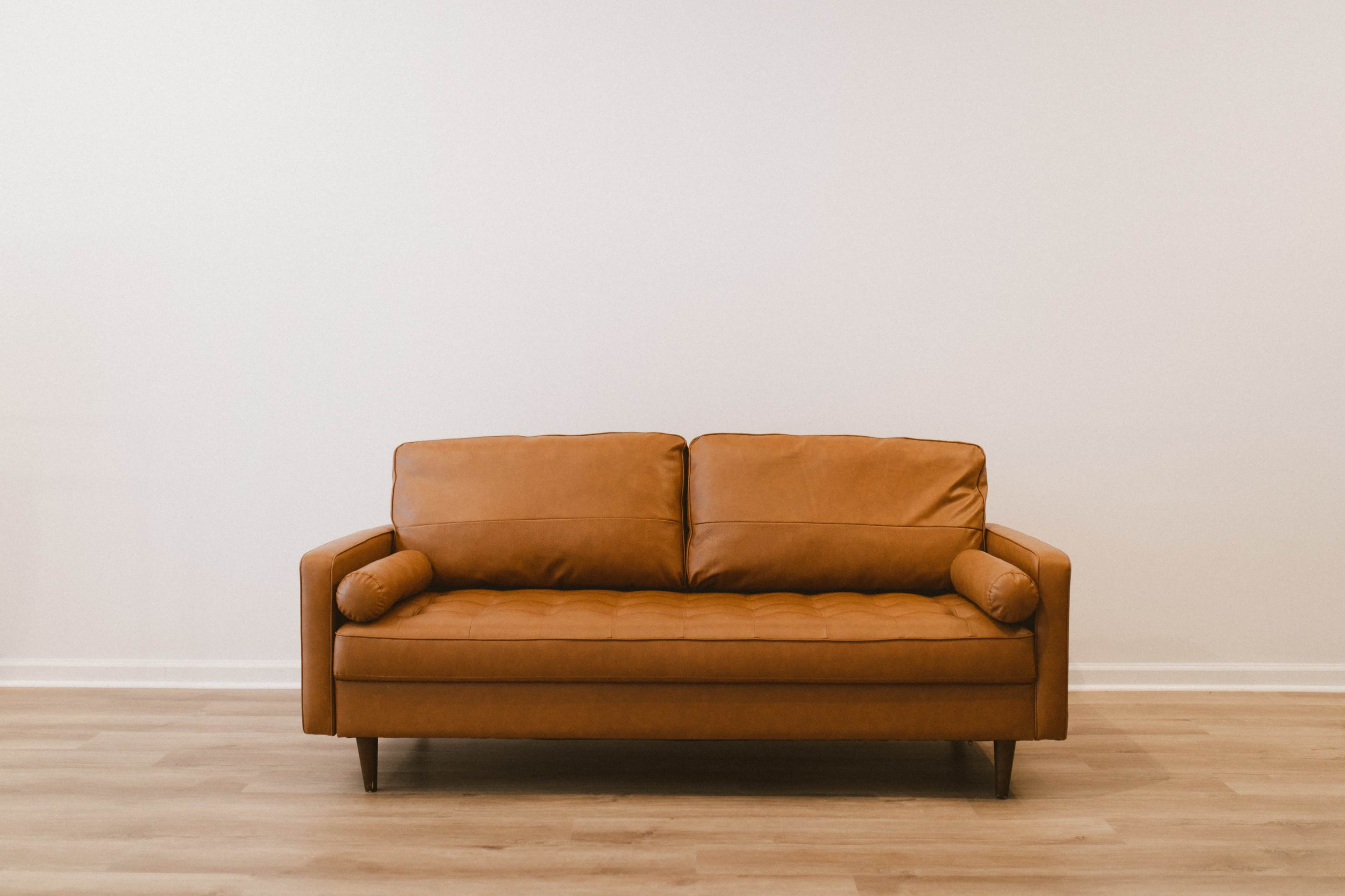 A brown leather couch with cylindrical arm cushions is positioned against a plain white wall.
