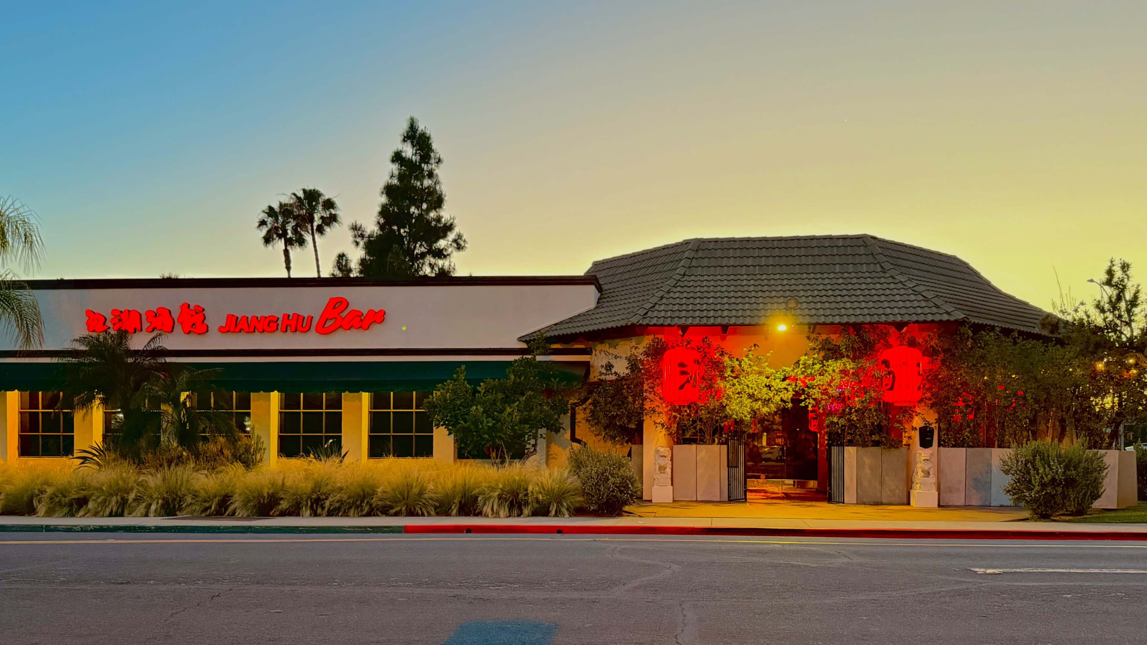 A restaurant with a thatched roof and illuminated red decorations stands beside a palm tree-lined road at sunset.