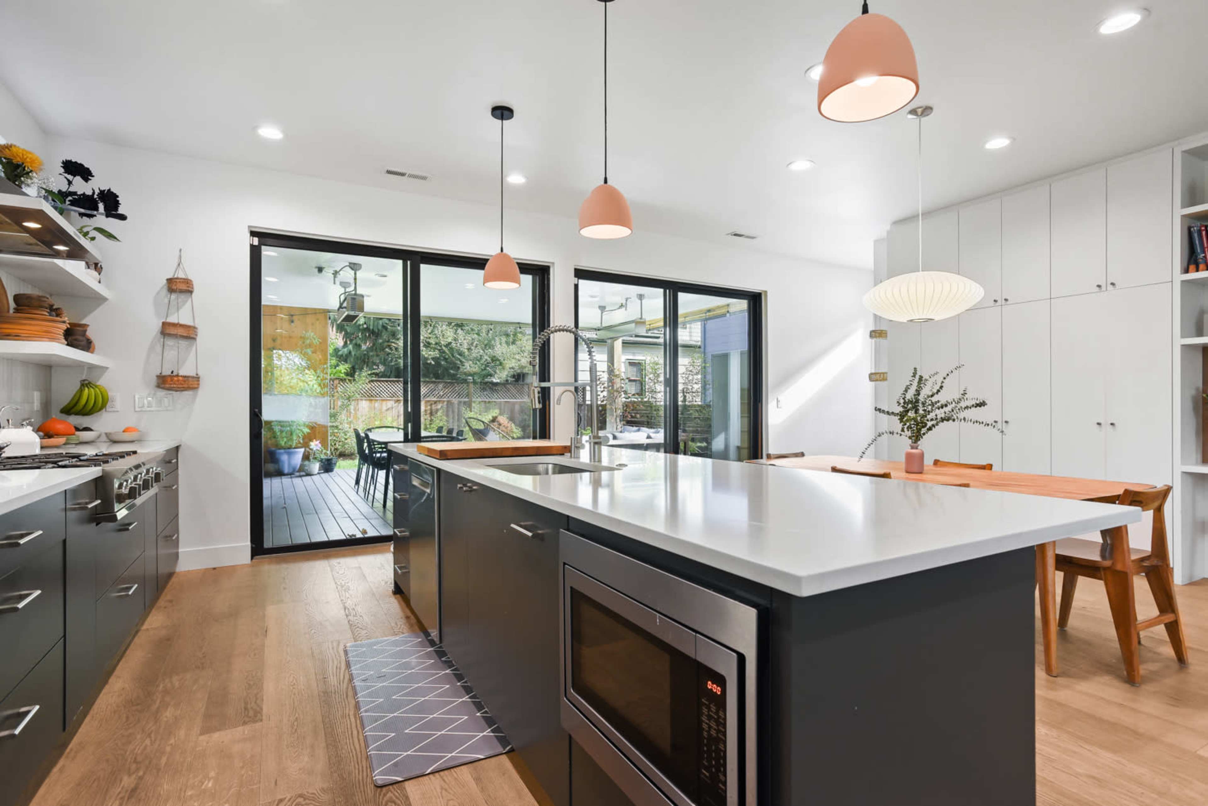 A modern kitchen features an island with a sink and microwave, light fixtures above, and large glass doors opening to an outdoor space.