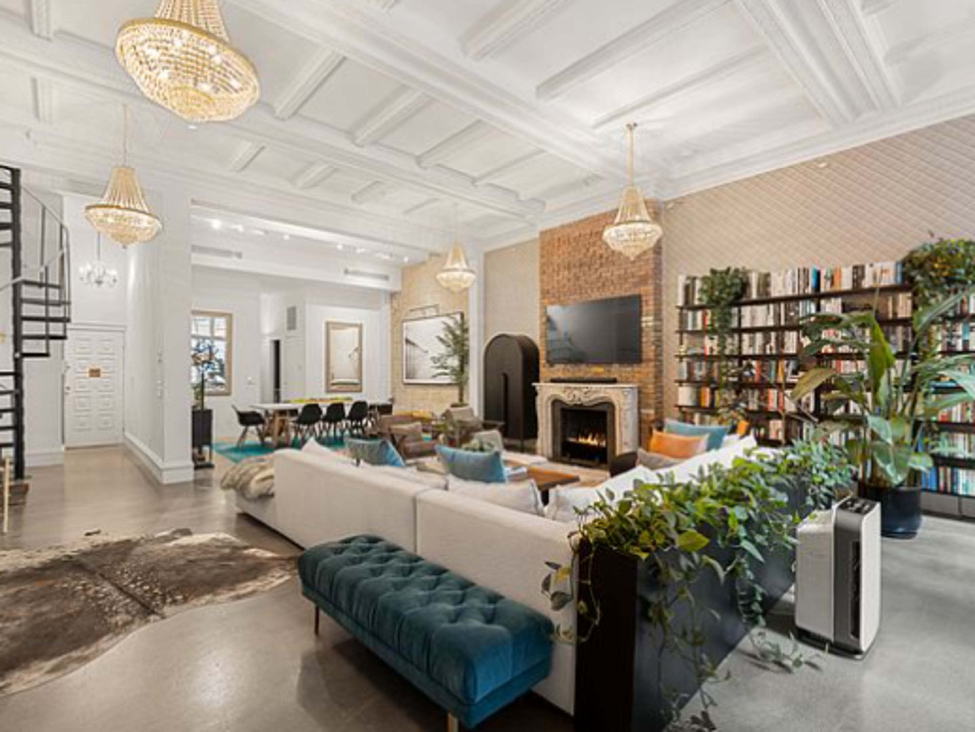 The image shows a spacious living room featuring a white sectional sofa, a blue velvet bench, a fireplace, and a large bookshelf filled with books, all illuminated by pendant chandeliers.