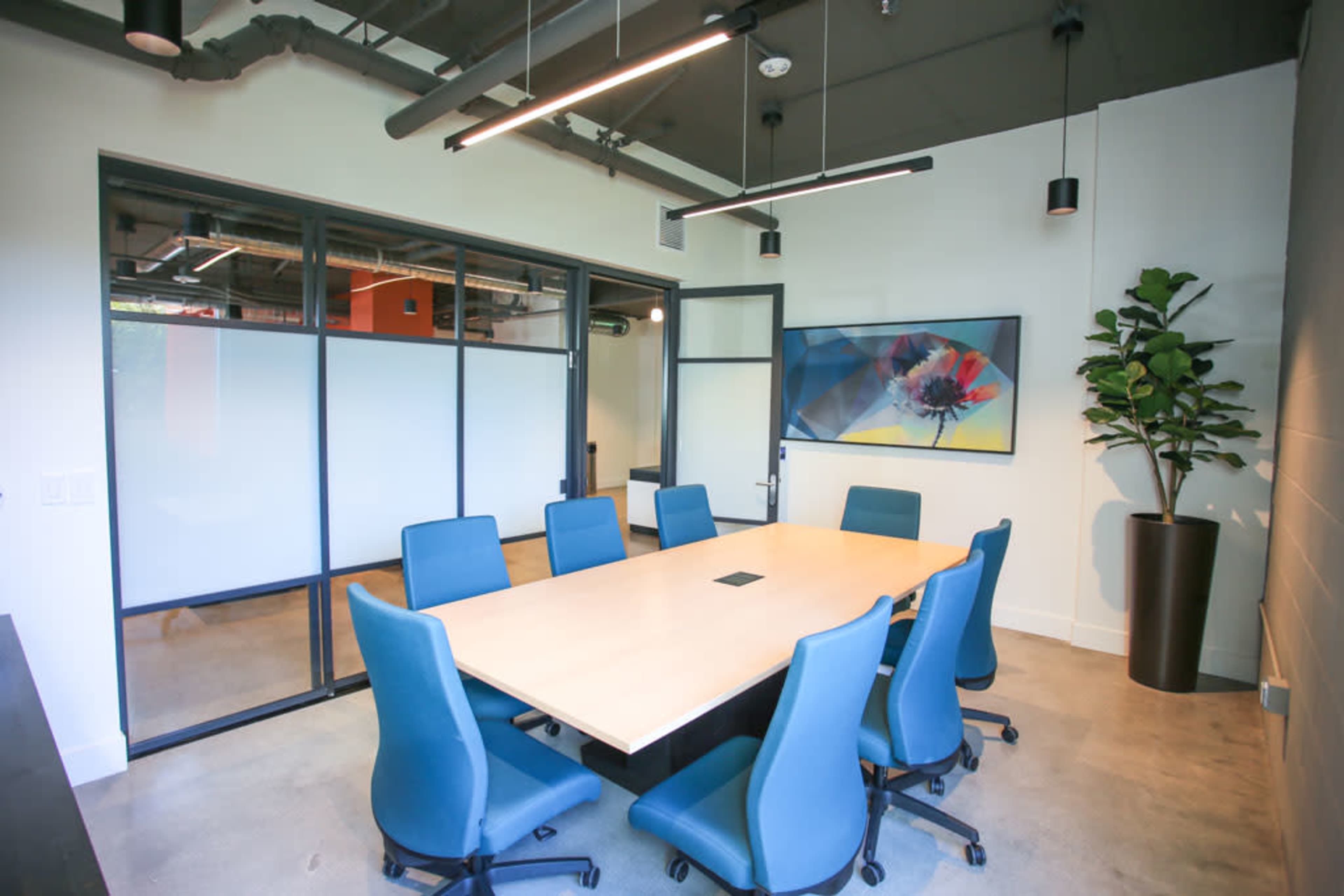 A modern conference room featuring a large rectangular table surrounded by blue chairs, with large windows and a potted plant.