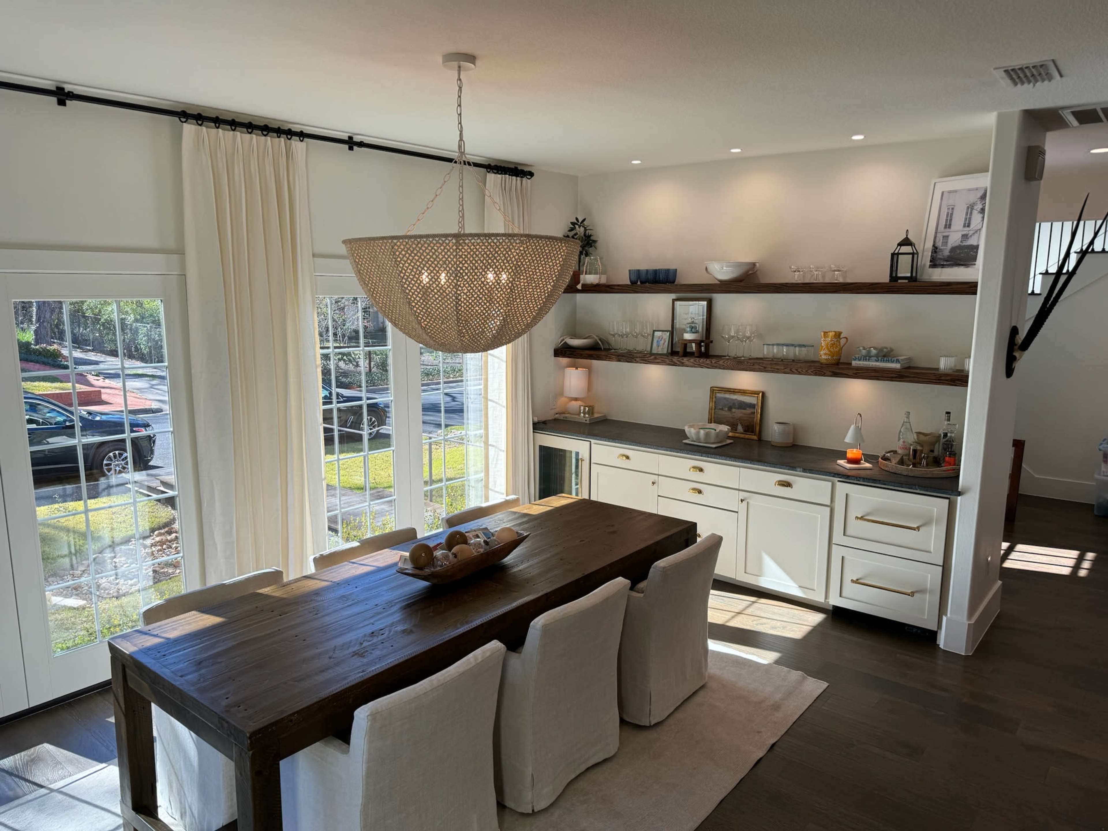 A modern dining area features a wooden table surrounded by upholstered chairs, with a large chandelier above and a sideboard filled with dishware.