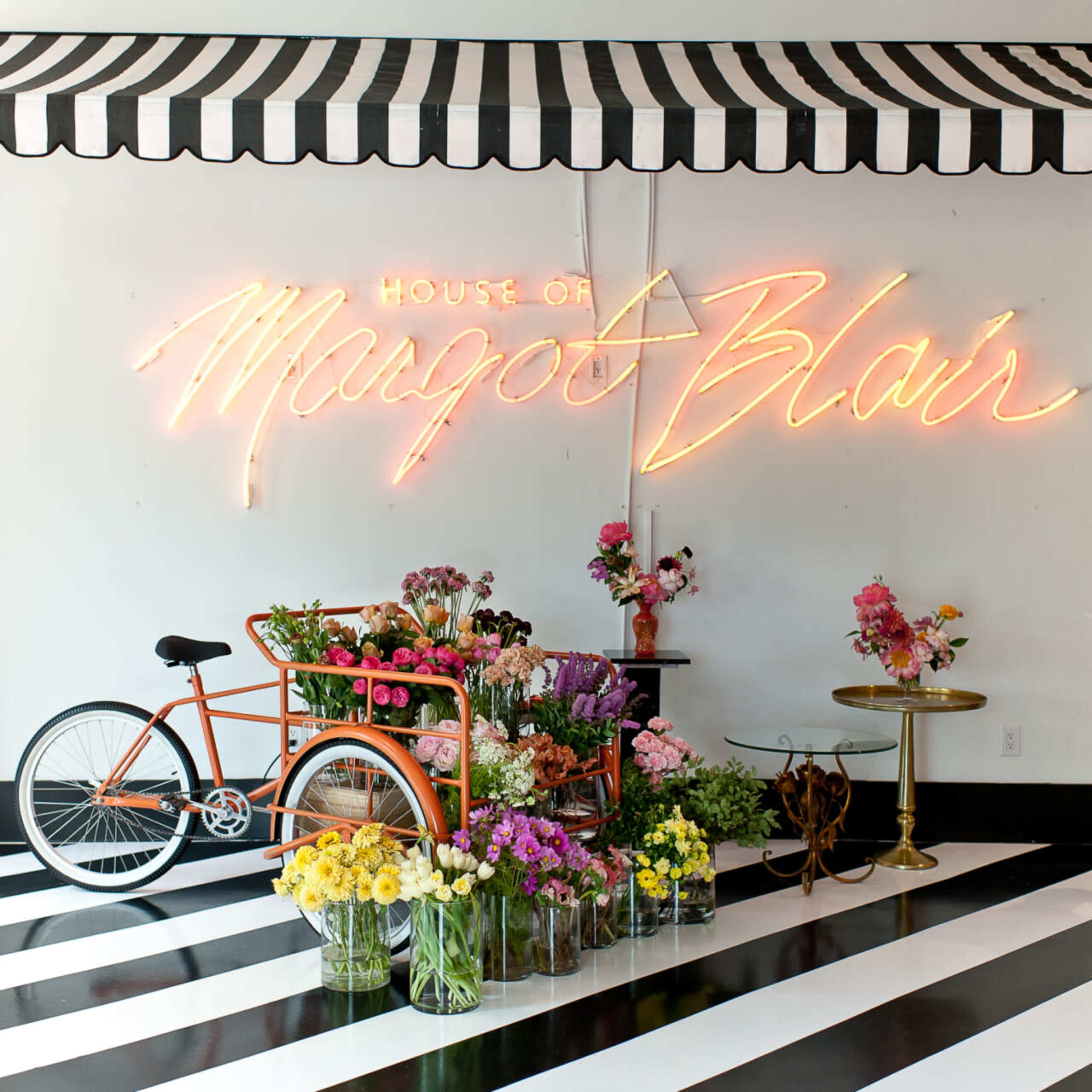 A brightly decorated flower shop features a neon sign reading "House of Margot Blair," an orange bicycle laden with flowers, and a black-and-white striped floor.