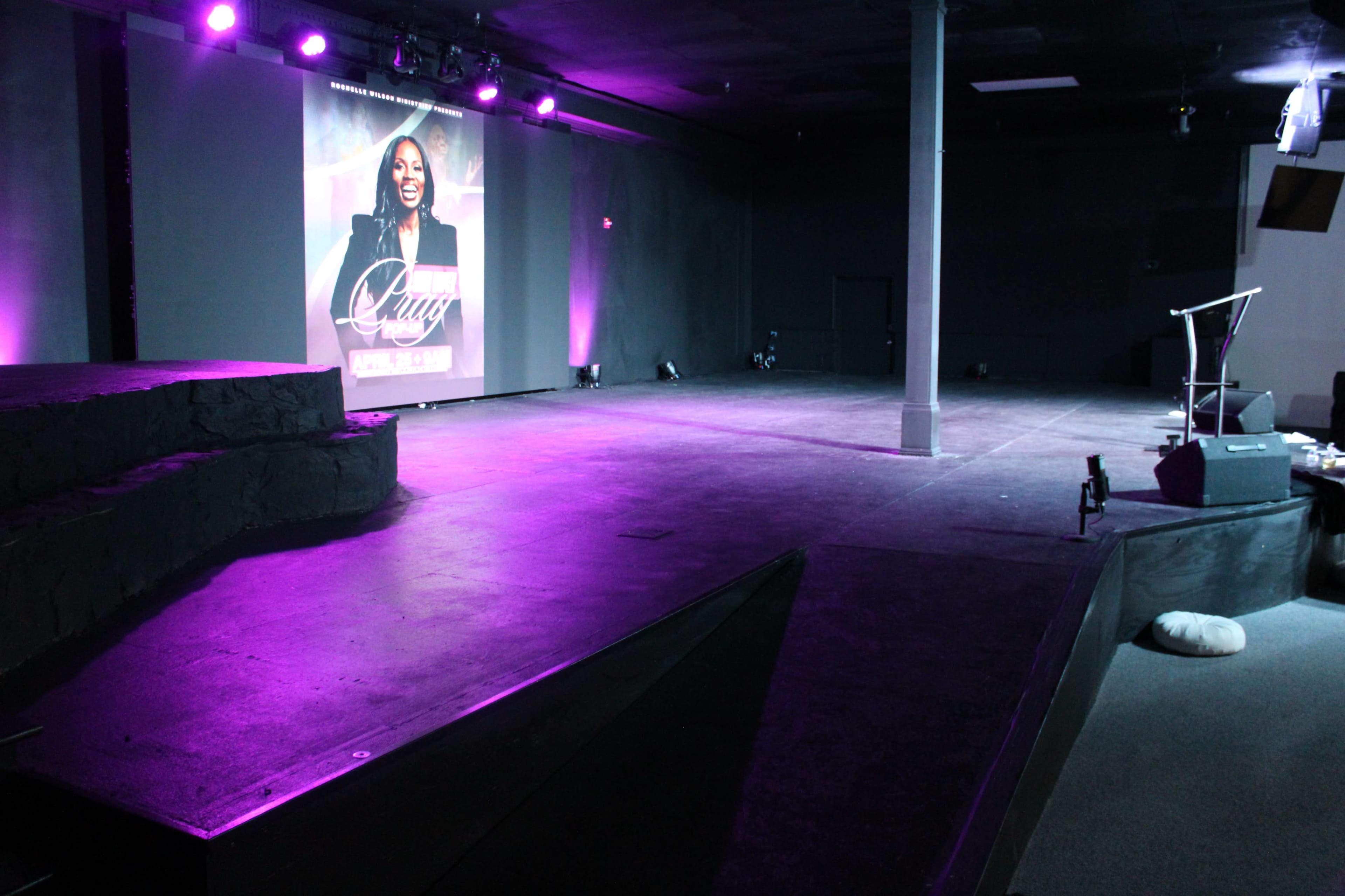 The image shows an empty stage with purple lighting, featuring a large promotional poster in the background and a microphone stand on the side.