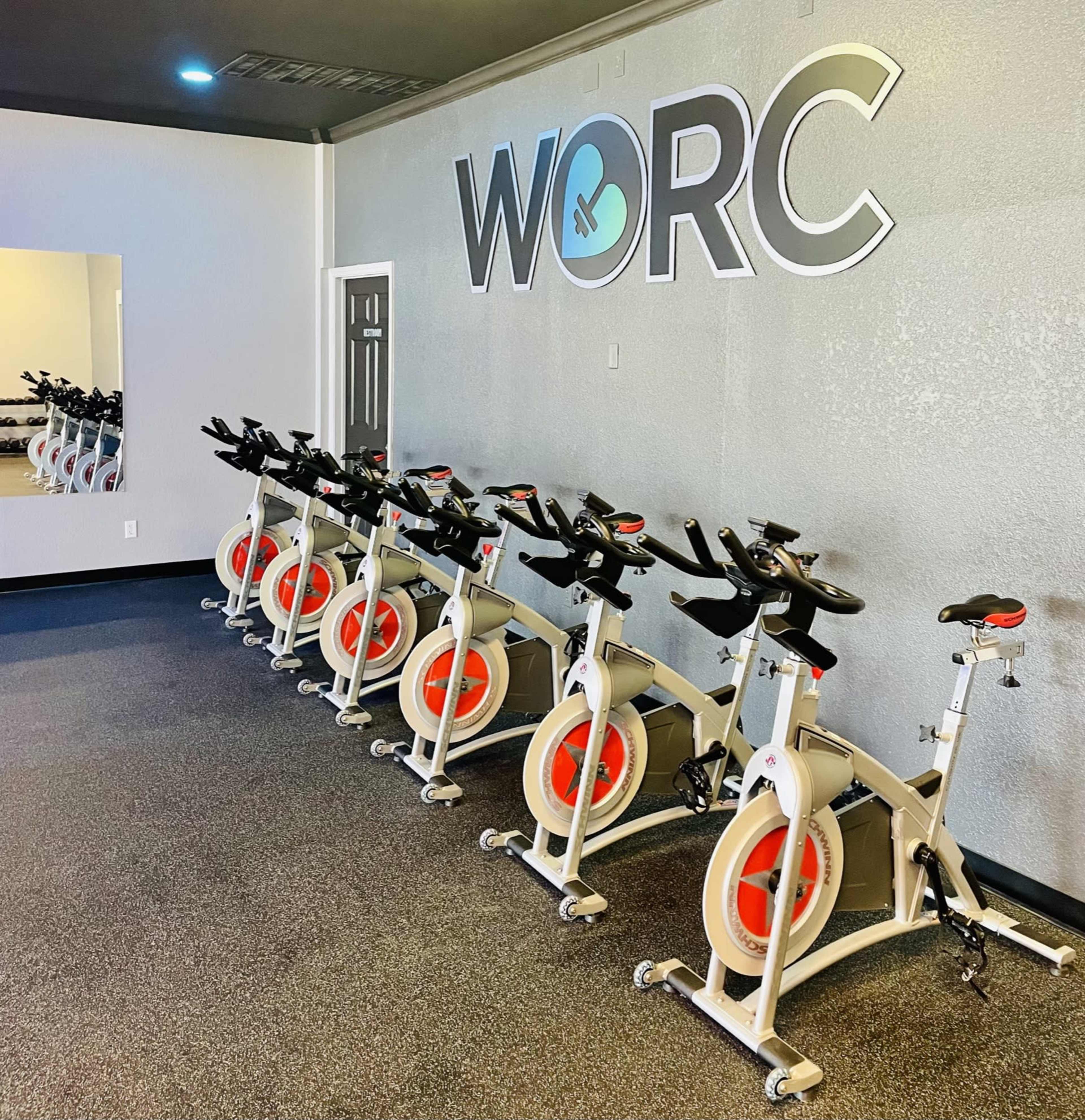 A row of stationary exercise bikes is lined up against a gray wall with the word "WORC" displayed prominently above them.