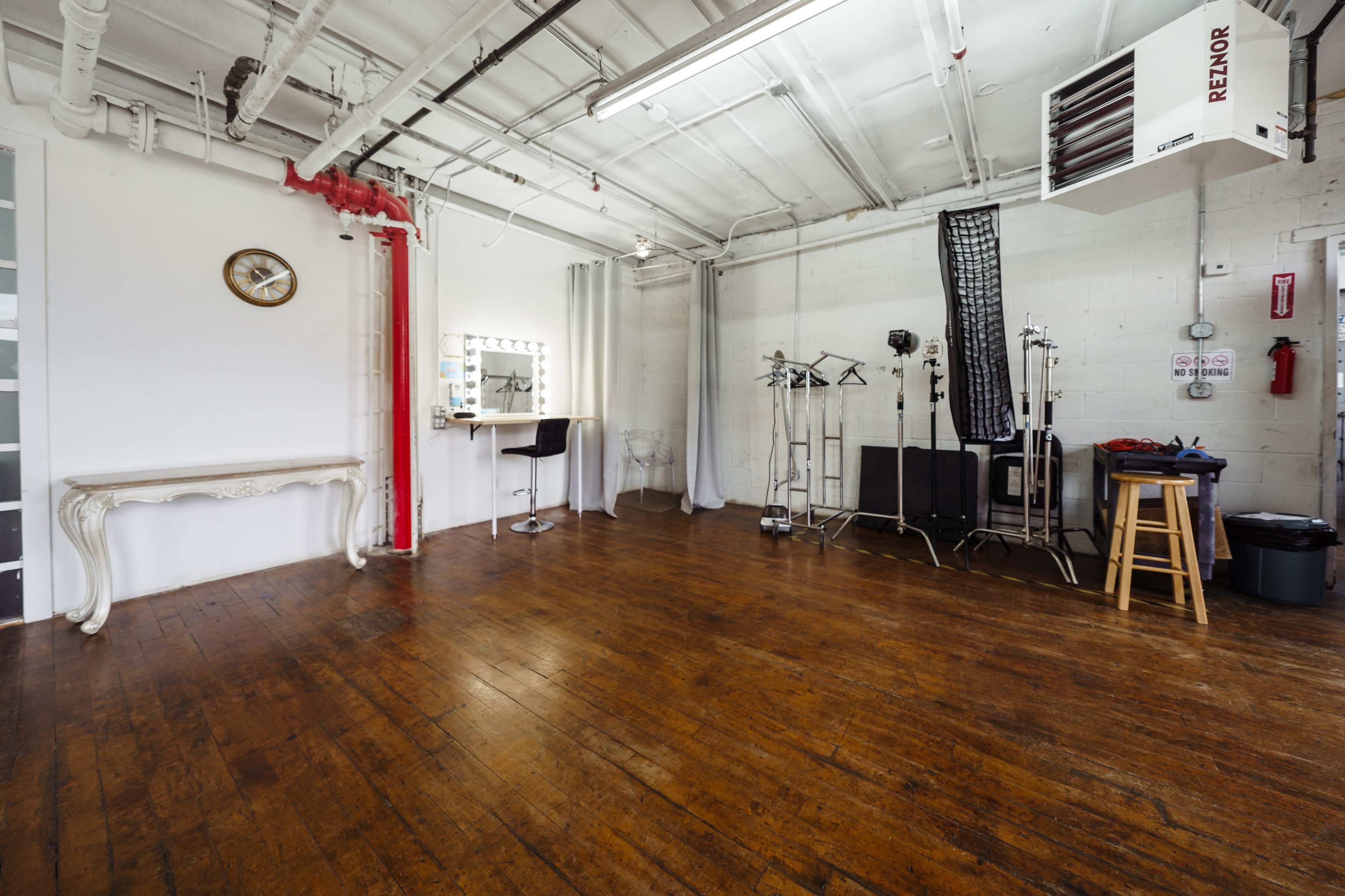 An empty studio space with wooden flooring, a large mirror, a clock on the wall, and various stands and equipment positioned around the room.