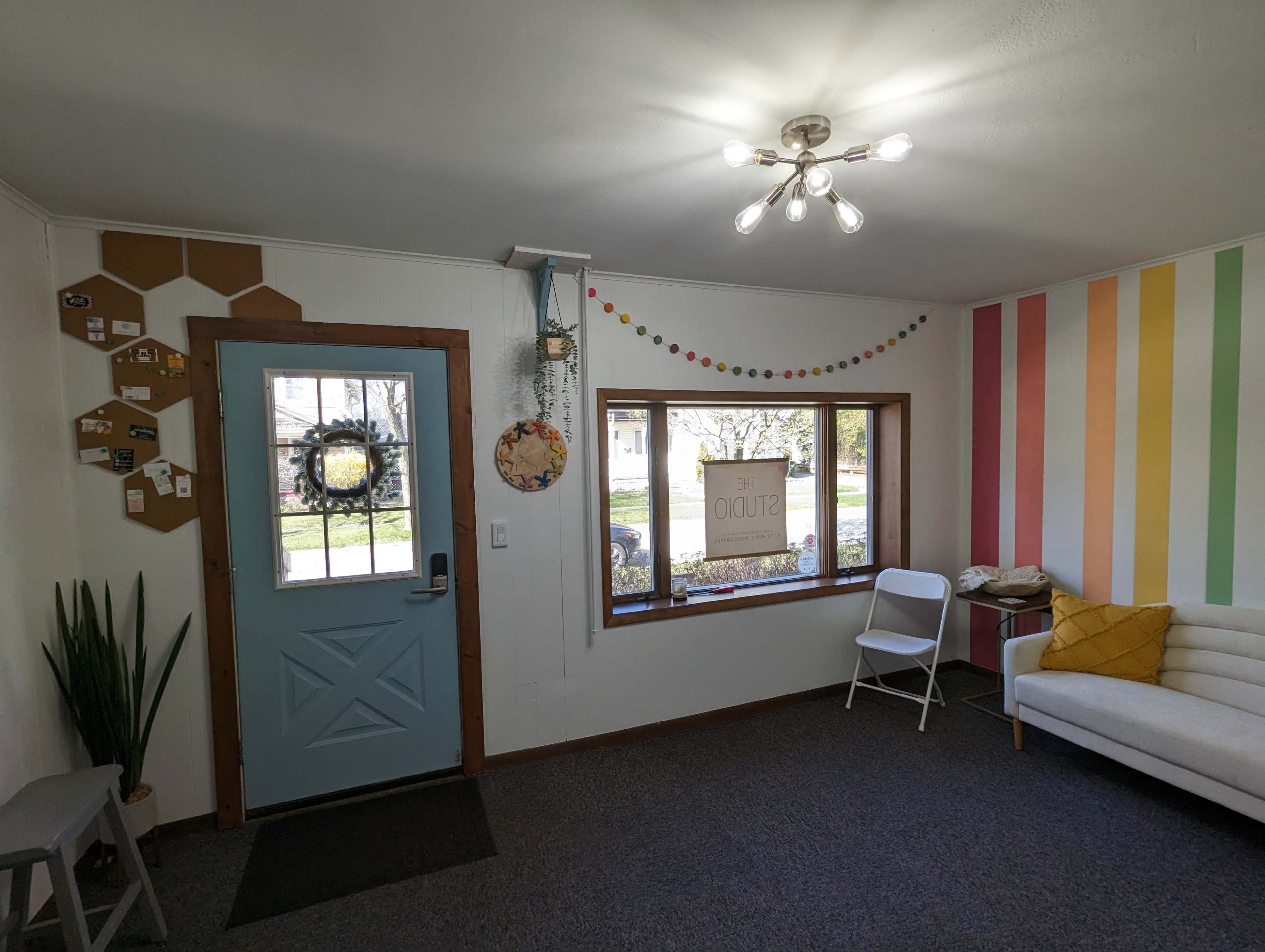 A cozy room with a blue door, a window featuring a hanging sign, and a colorful striped wall.
