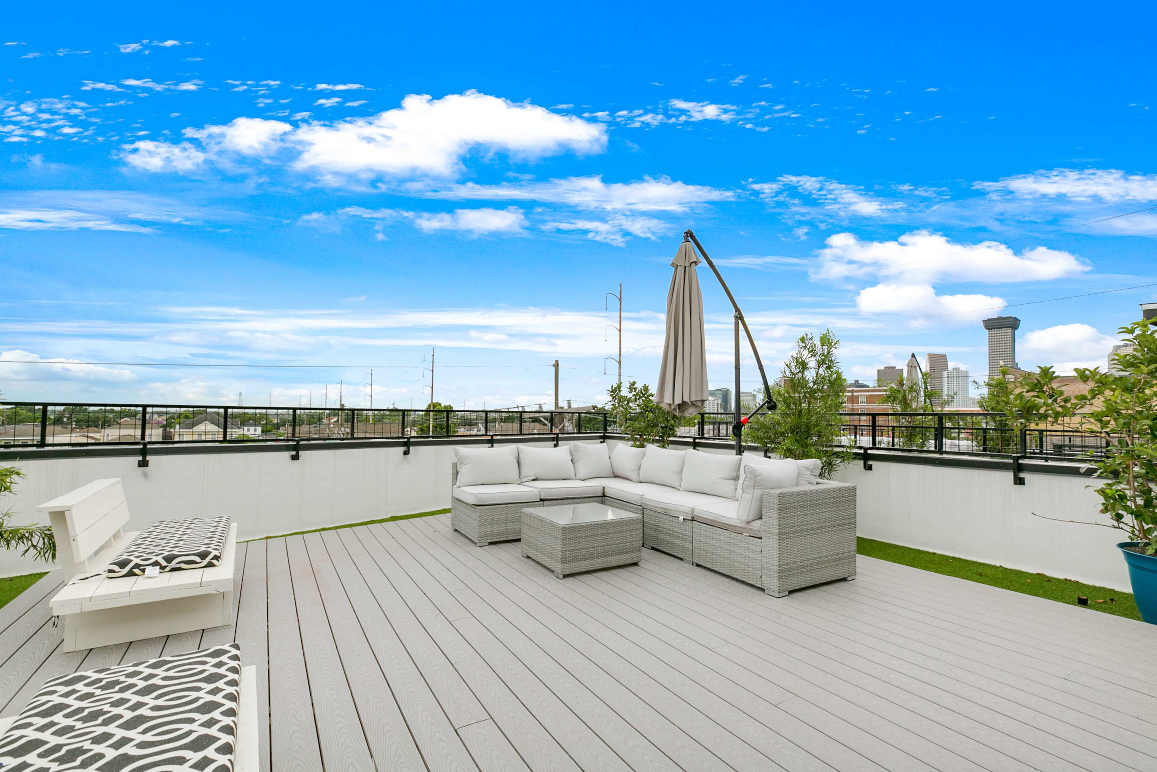 A rooftop deck with a seating area, umbrella, and city skyline in the background under a clear blue sky.