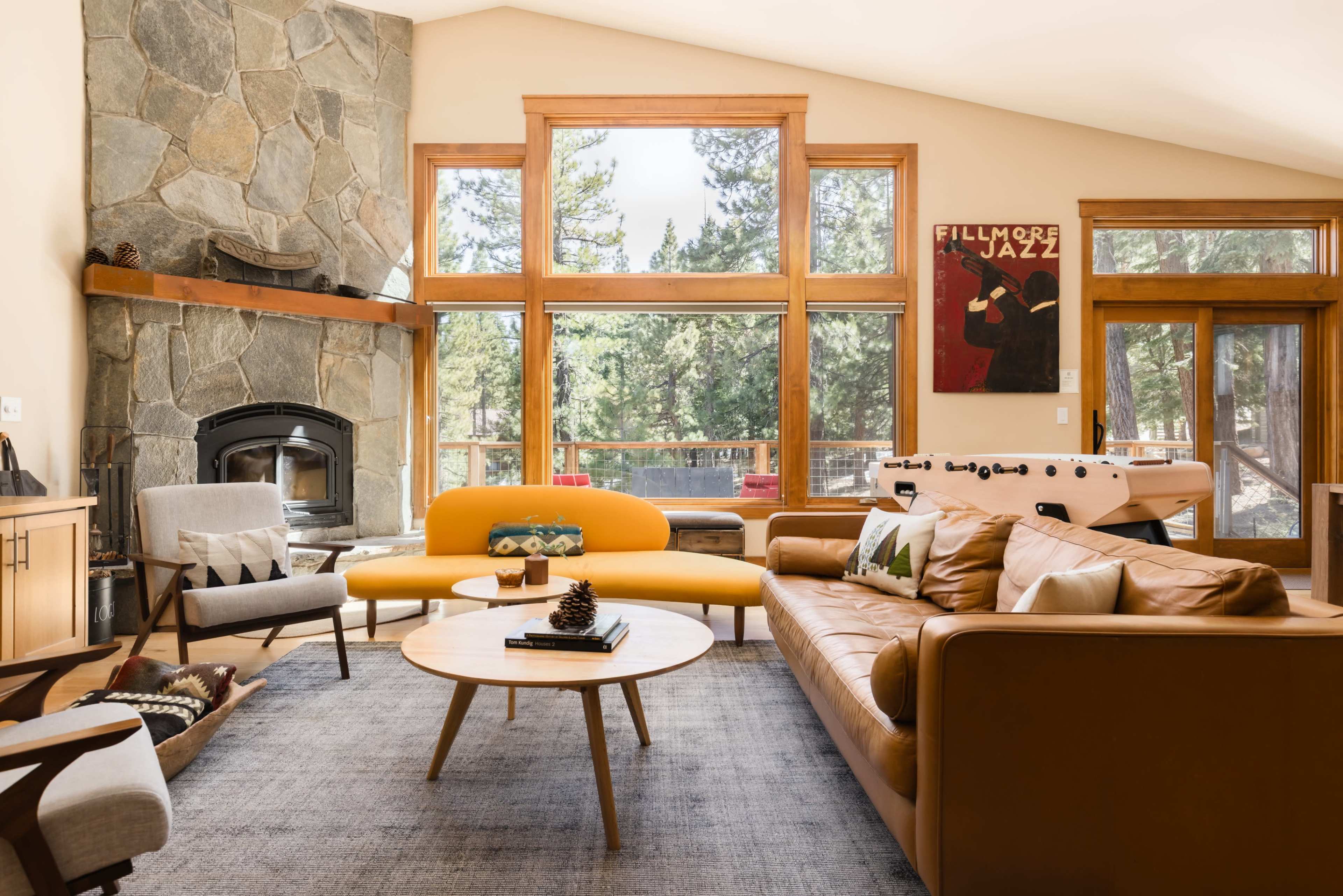 A spacious living room features a stone fireplace, large windows overlooking trees, and a mix of modern furniture, including a yellow sofa and a coffee table.