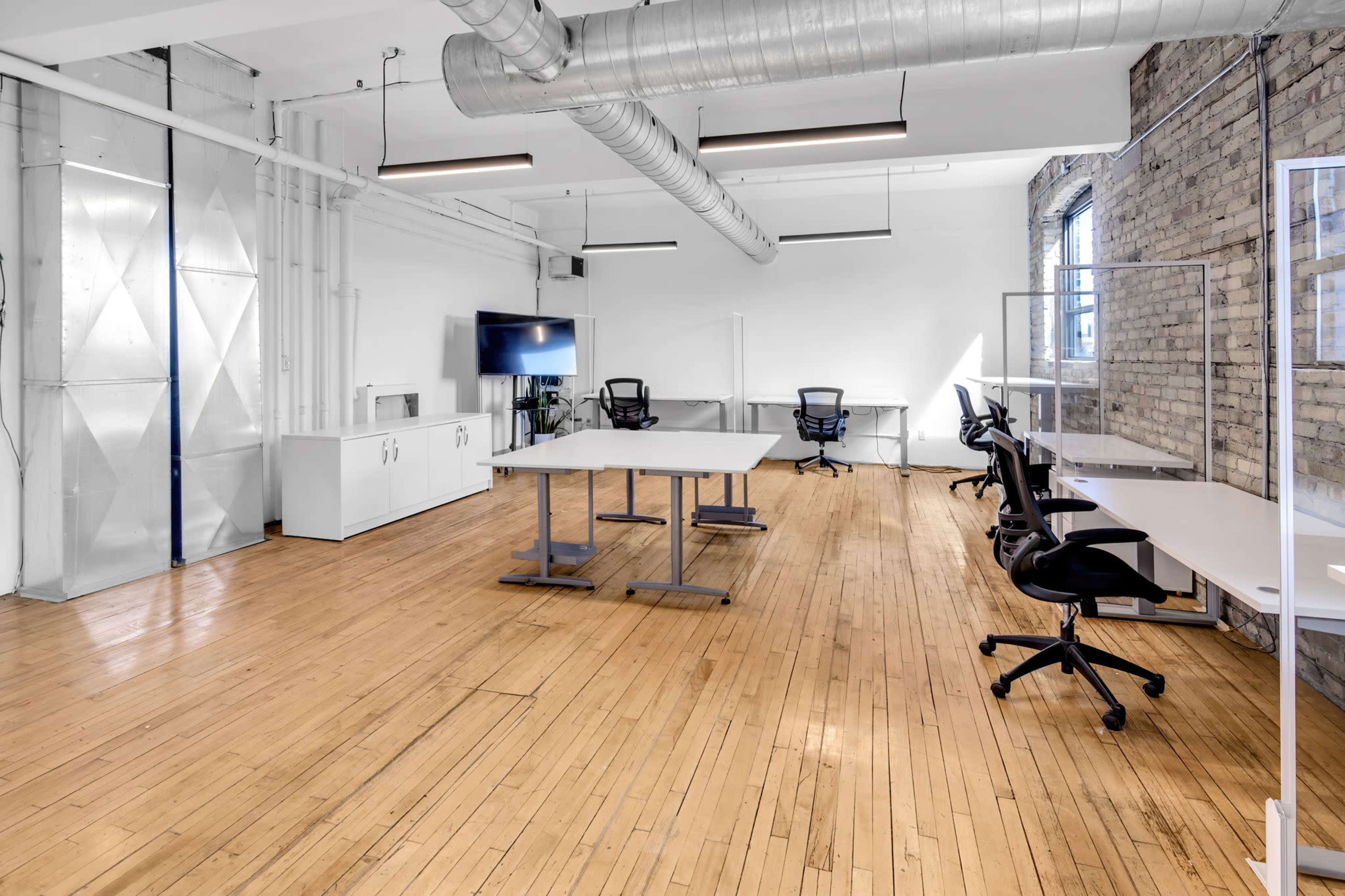 The image shows a modern office space with hardwood floors, featuring several desks and chairs, a large screen, and glass partitions.