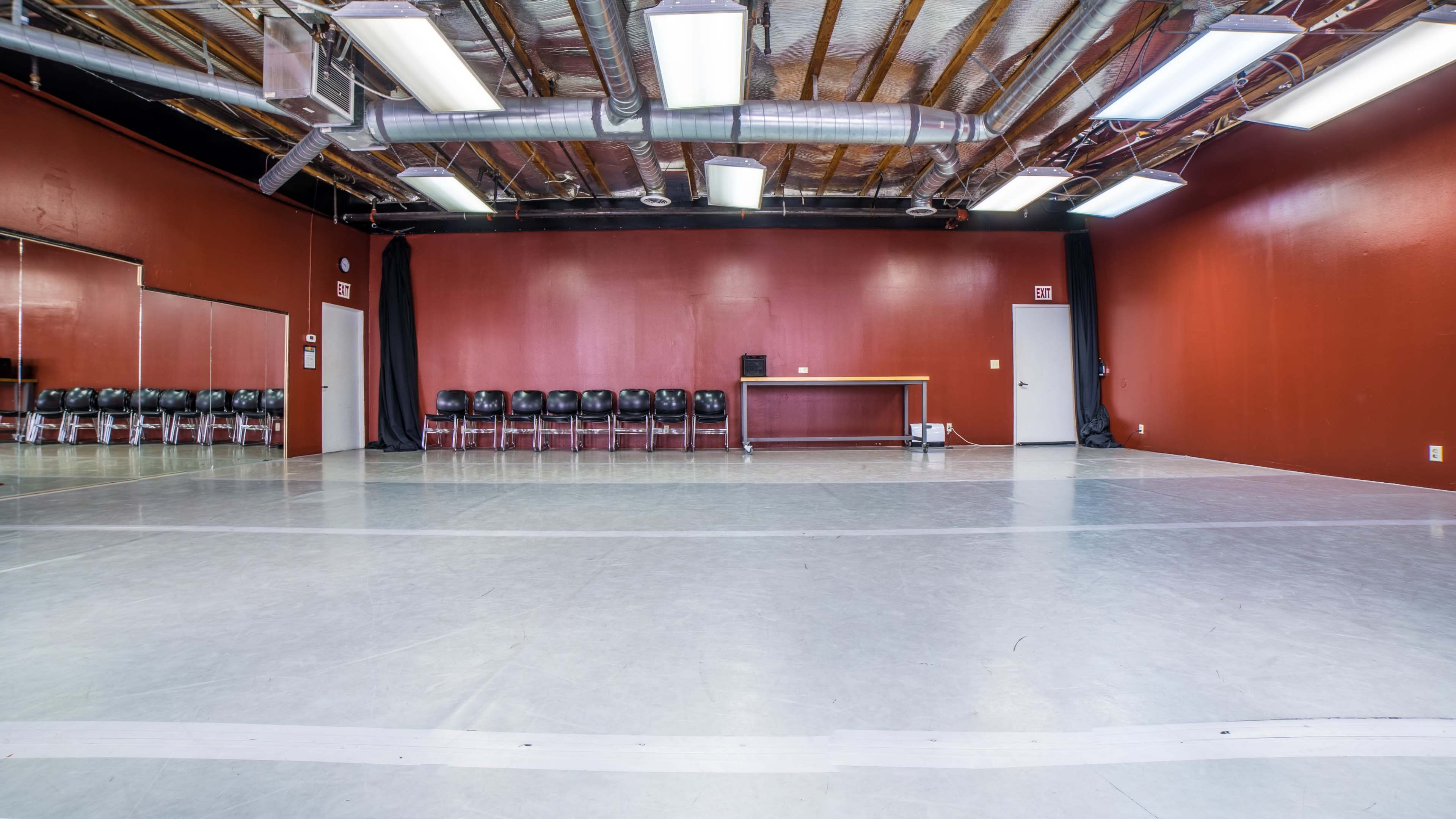 A spacious dance studio with a gray floor, red walls, and a row of black chairs along one side.
