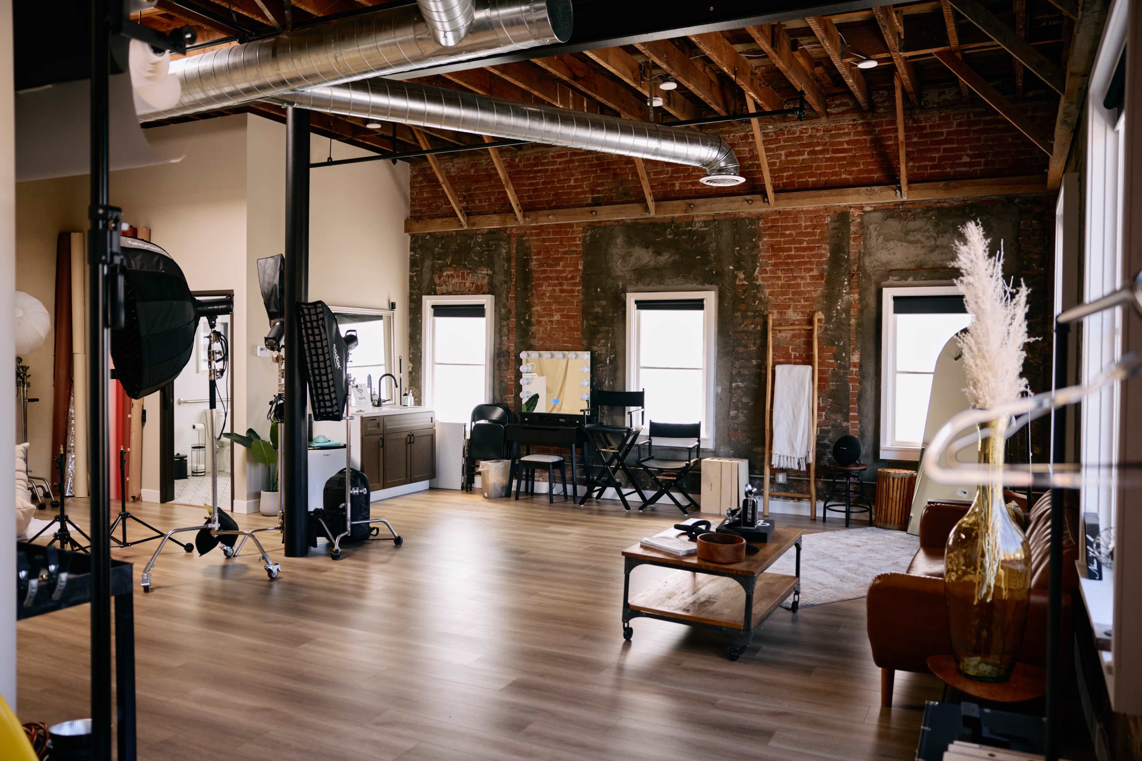 A spacious studio with exposed brick walls, wooden beams, and a mixture of furniture including a couch, a table, and black director's chairs.