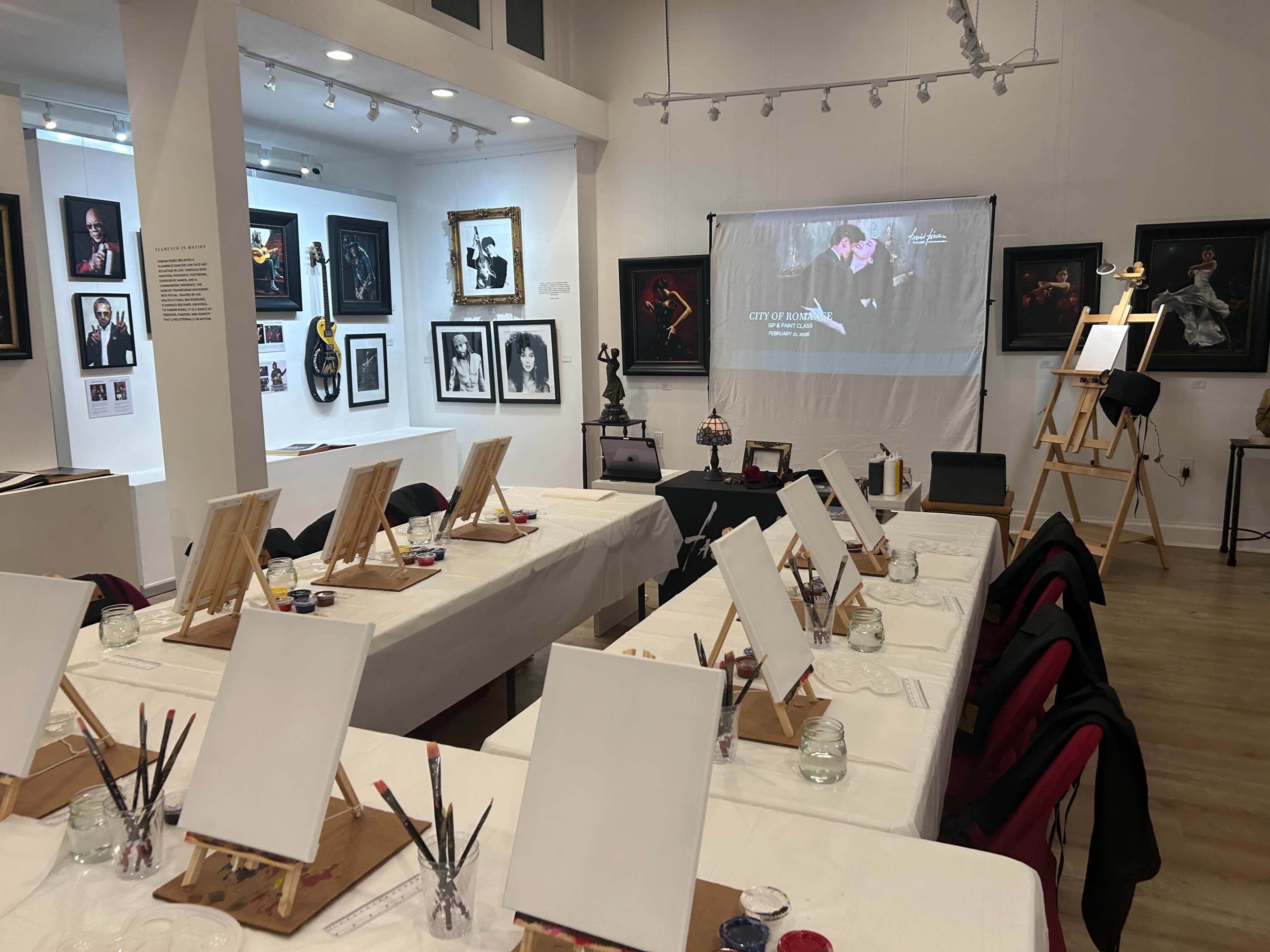 The image shows an art studio with several tables set up for painting, each equipped with canvases, paint supplies, and brushes, and featuring various artworks displayed on the walls.