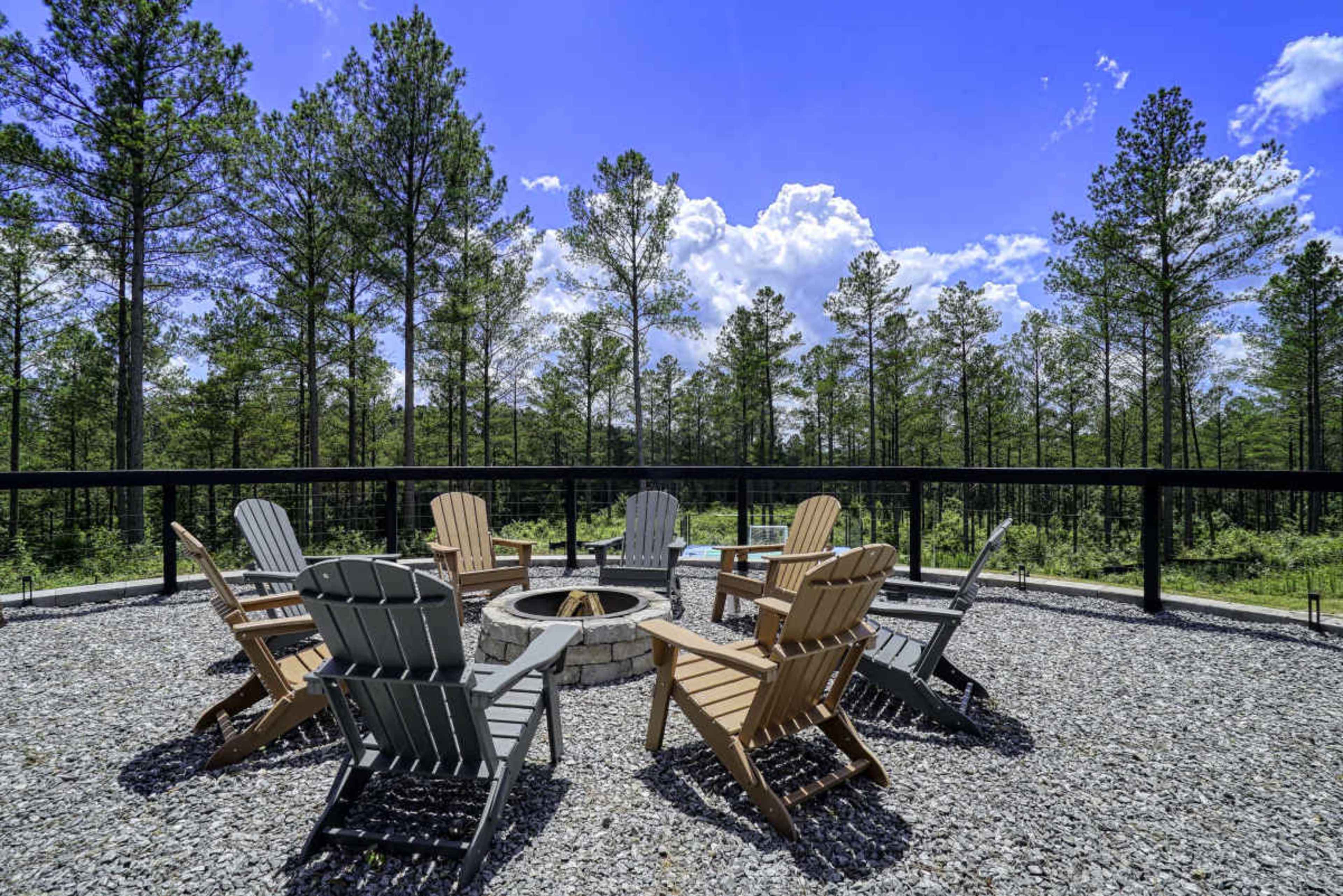 A circular fire pit surrounded by several wooden adirondack chairs sits within a gravel area, framed by tall trees and a clear blue sky.