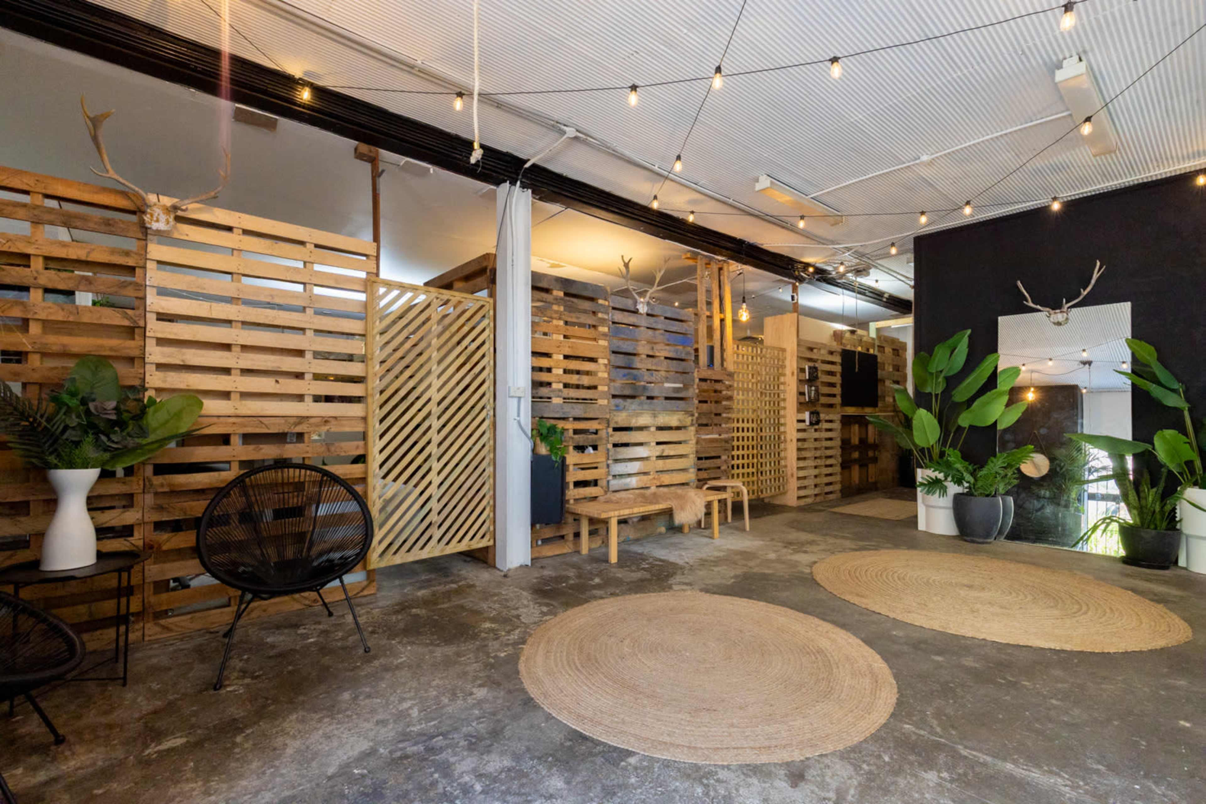 The image shows a modern indoor space with wooden slat partitions, circular rugs on a concrete floor, and potted plants placed around black seating.