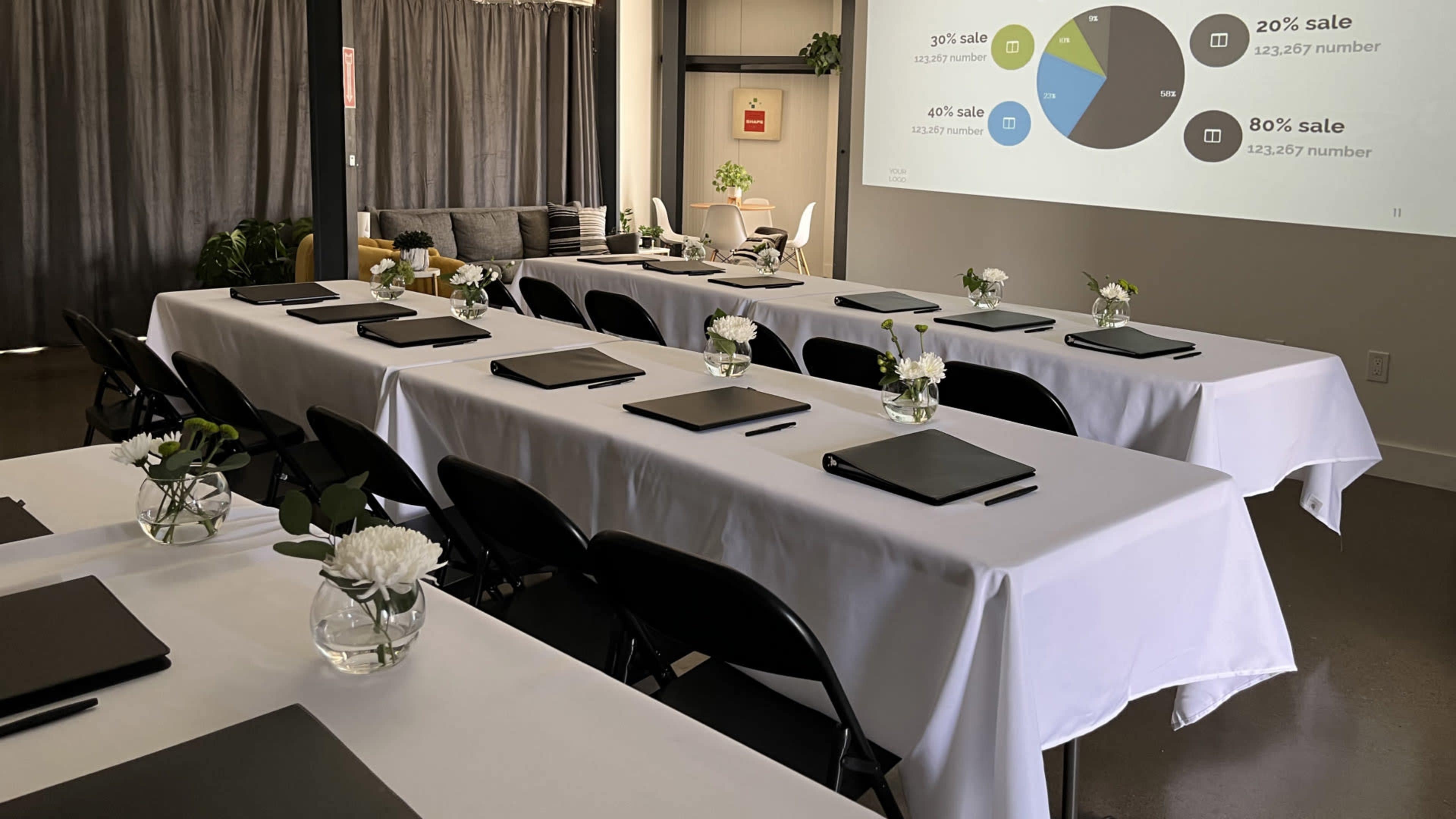 A conference room is set up with neatly arranged tables covered in white tablecloths, each equipped with laptops and small vases of flowers, while a presentation screen displays pie charts and sale percentages.