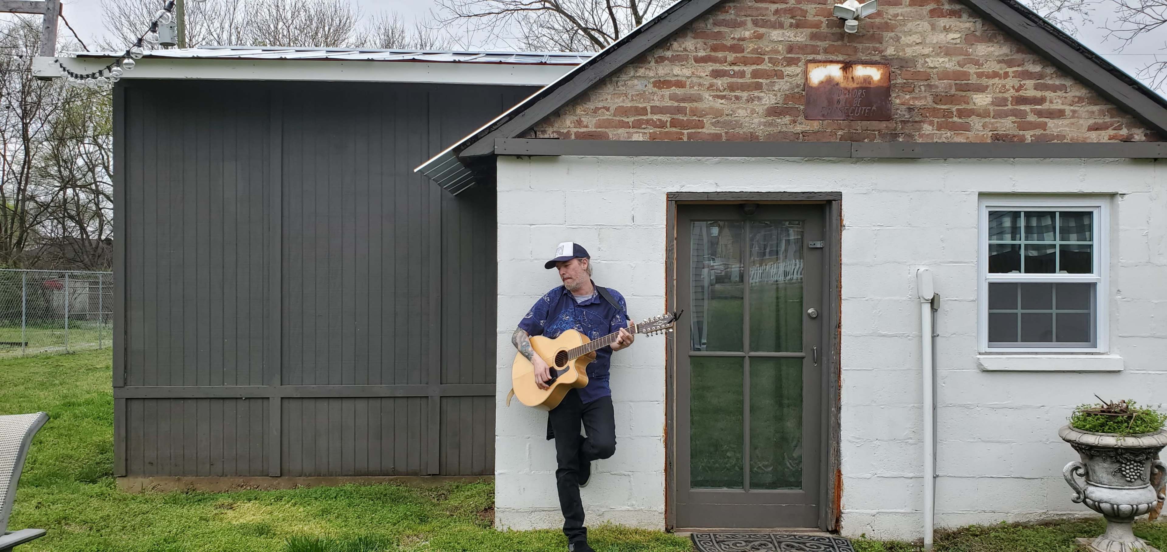 A man stands against a white brick building while playing an acoustic guitar in a grassy yard.