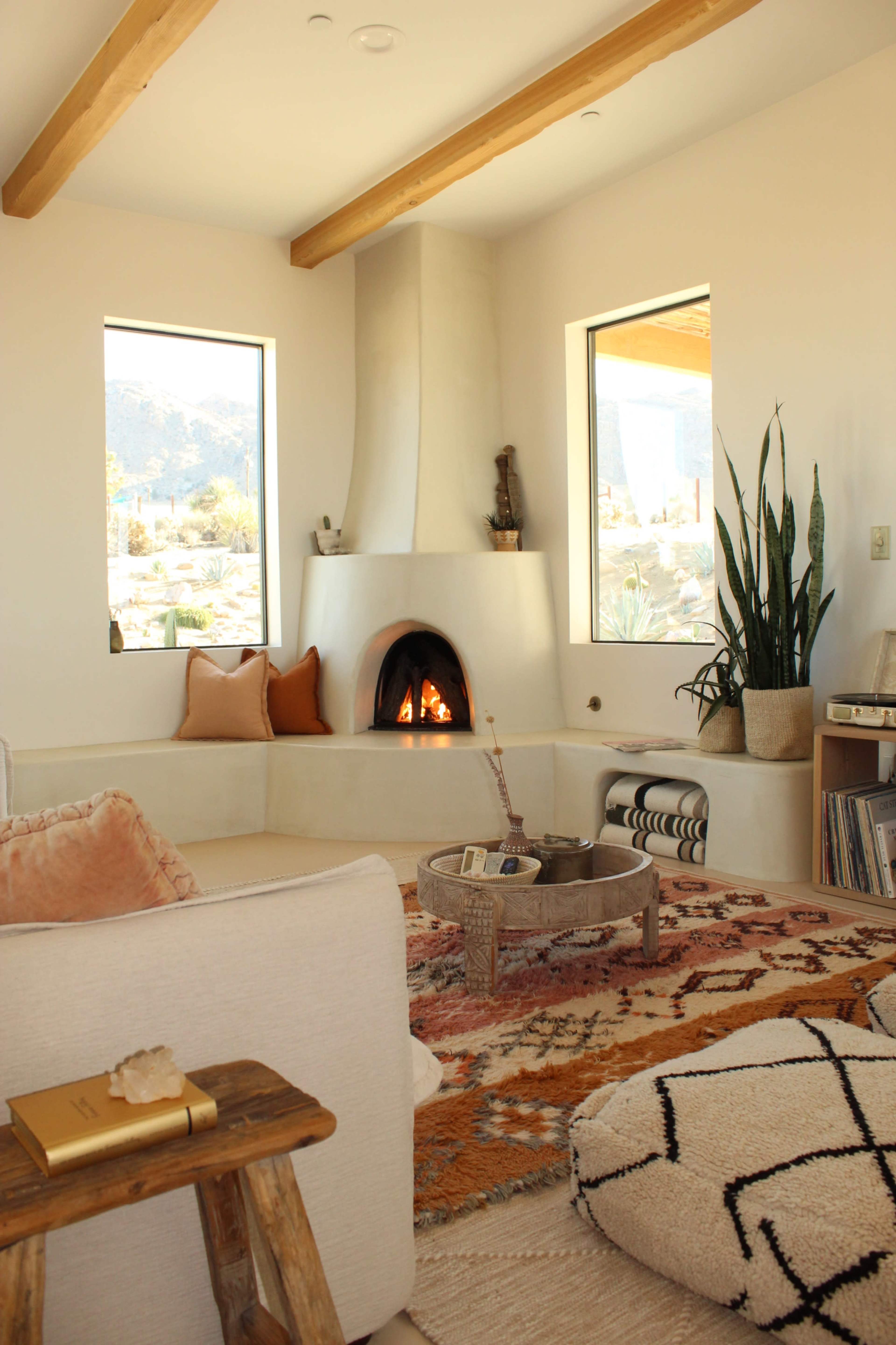 A cozy living room features a fireplace at the center, surrounded by windows showcasing a mountainous landscape, with a comfortable seating area and decorative plants.