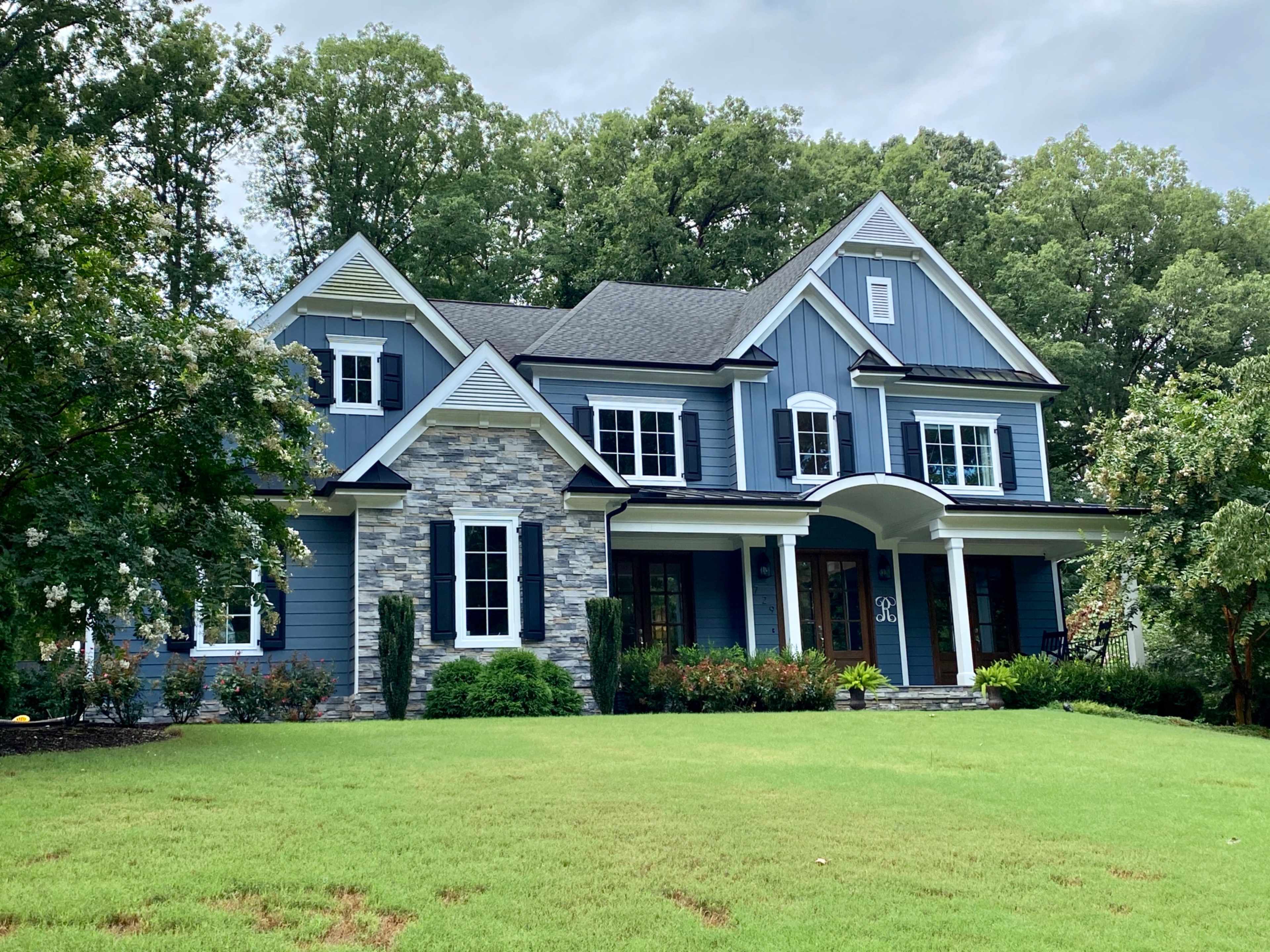 A large blue home with stone accents and multiple gables sits amidst a well-manicured lawn and trees.