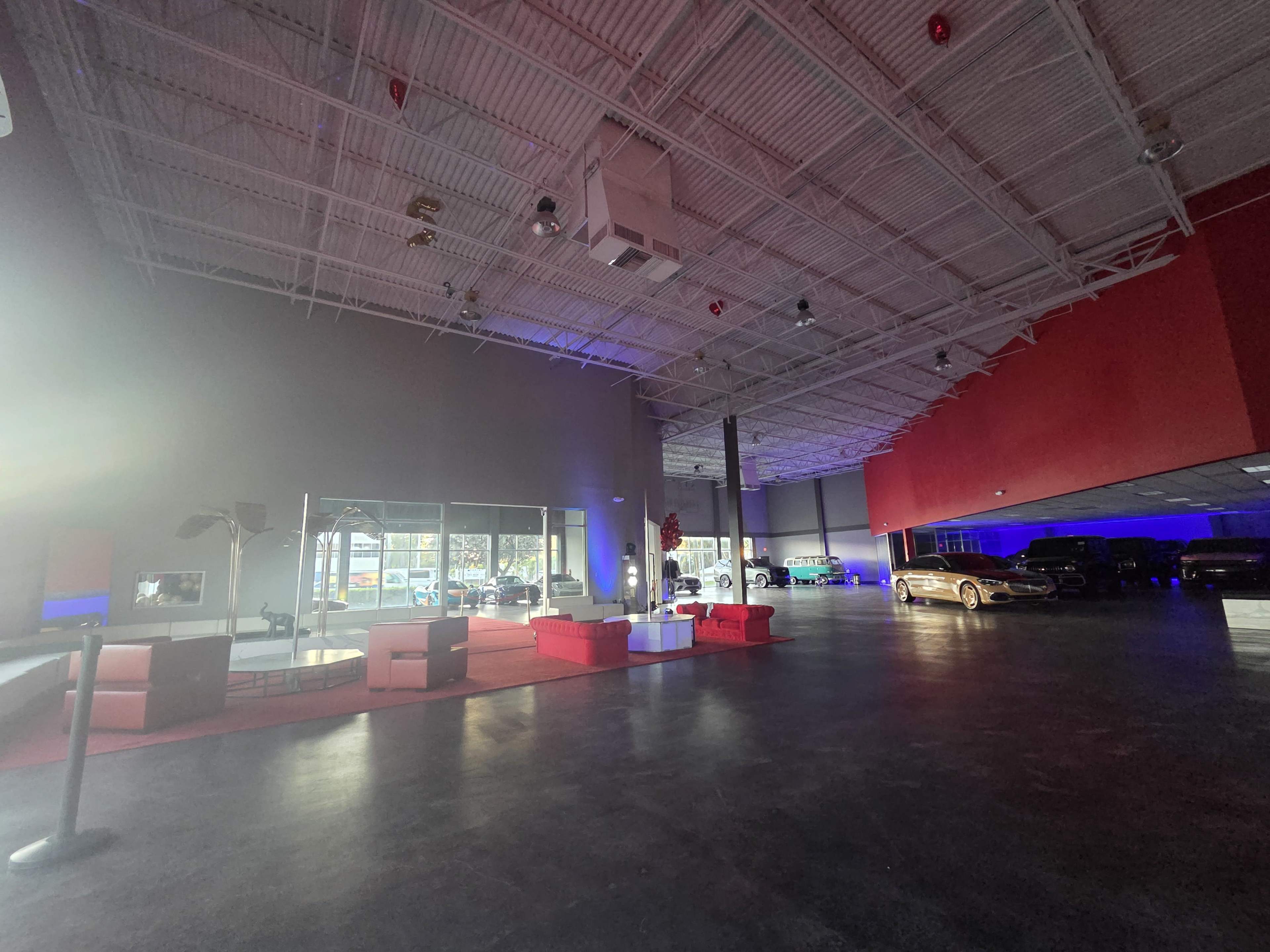 The image shows a spacious car showroom with various classic cars displayed under bright lighting and a modern seating area.