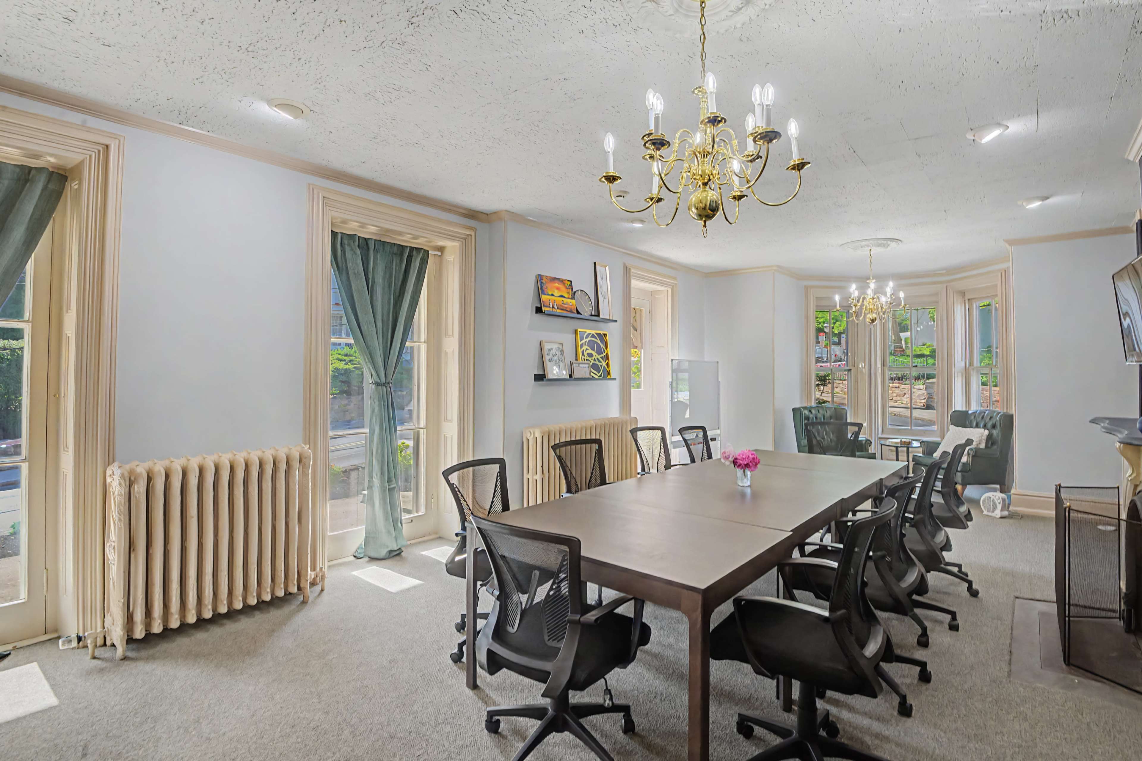 A spacious conference room features a large wooden table surrounded by black office chairs, with tall windows letting in natural light and a chandelier hanging from the ceiling.