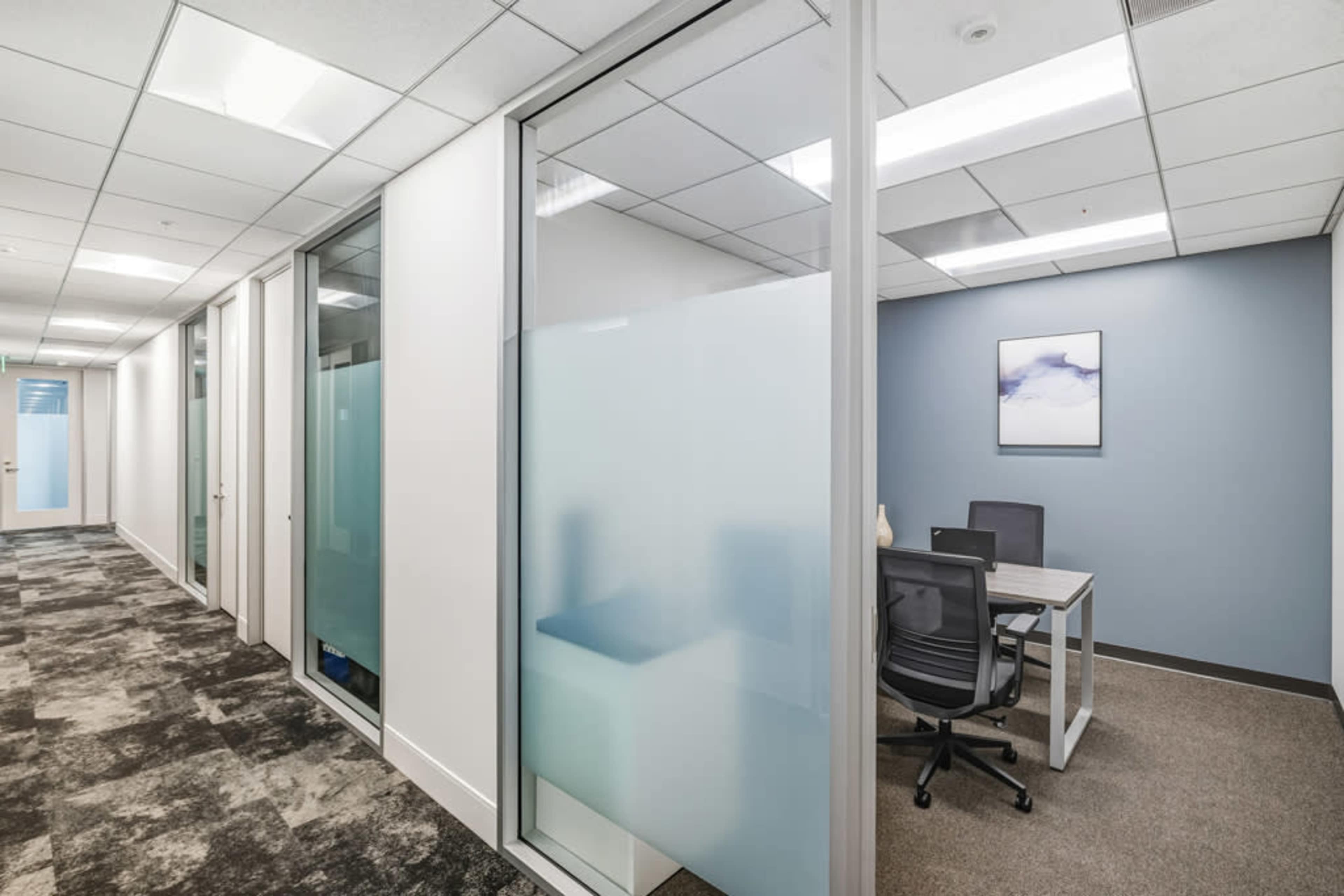 The image shows a modern office corridor with frosted glass-walled meeting rooms and a workspace featuring a desk and chair.