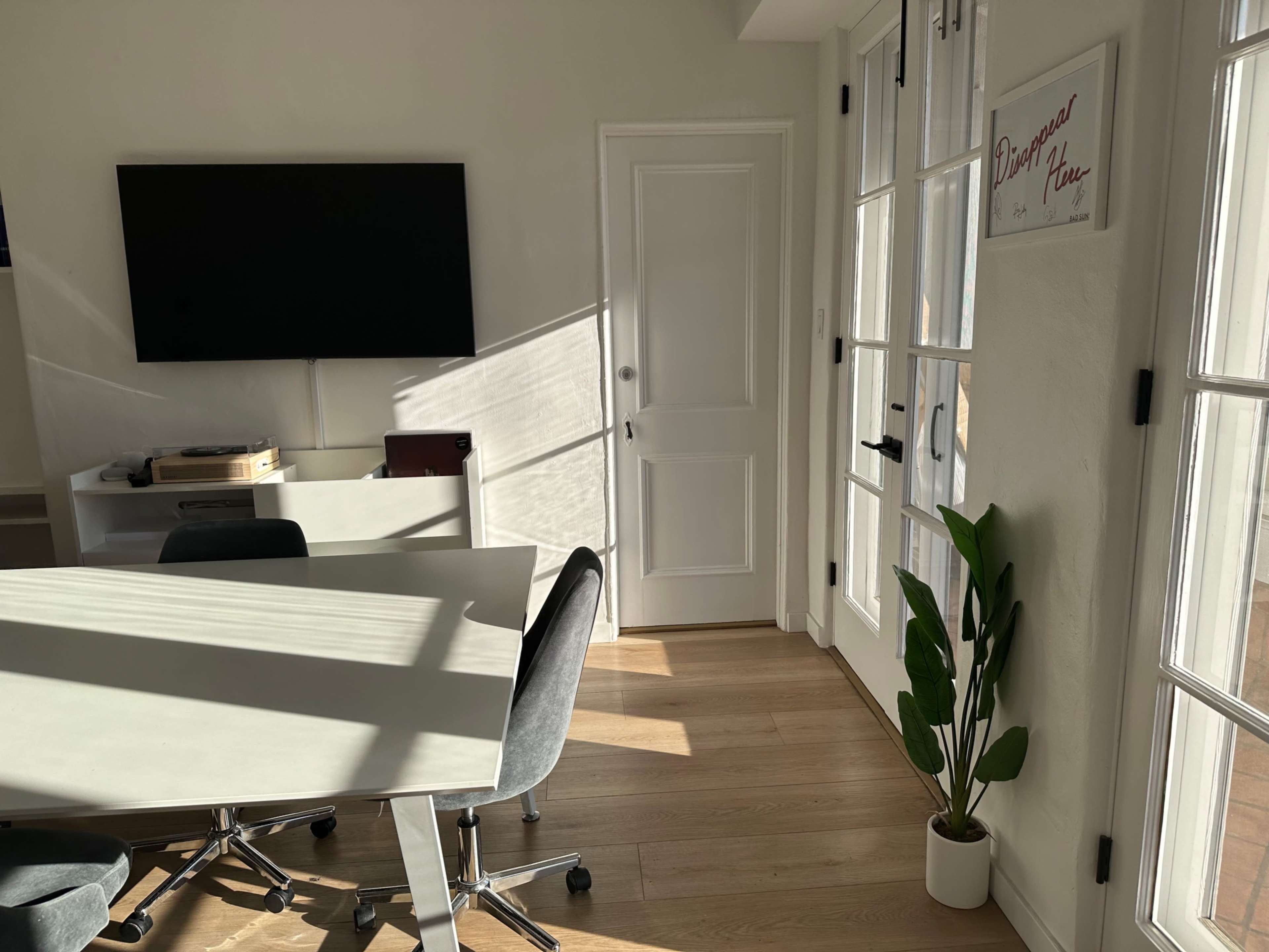 A modern office space features a table with chairs, a wall-mounted screen, large windows allowing sunlight to stream in, and a potted plant in the corner.