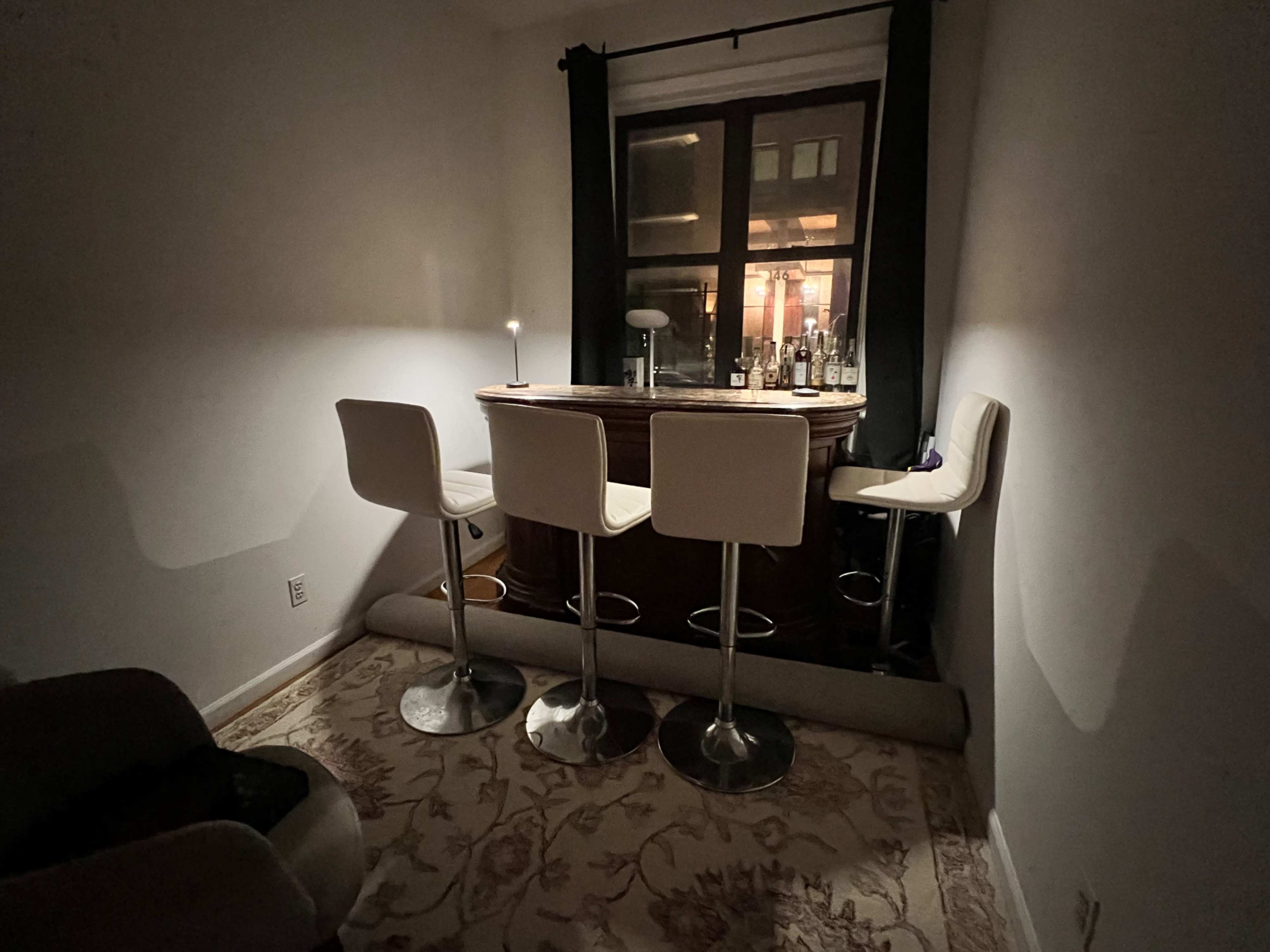 A small home bar area with three high stools and a wooden counter, positioned near a window displaying bottles on the shelf.