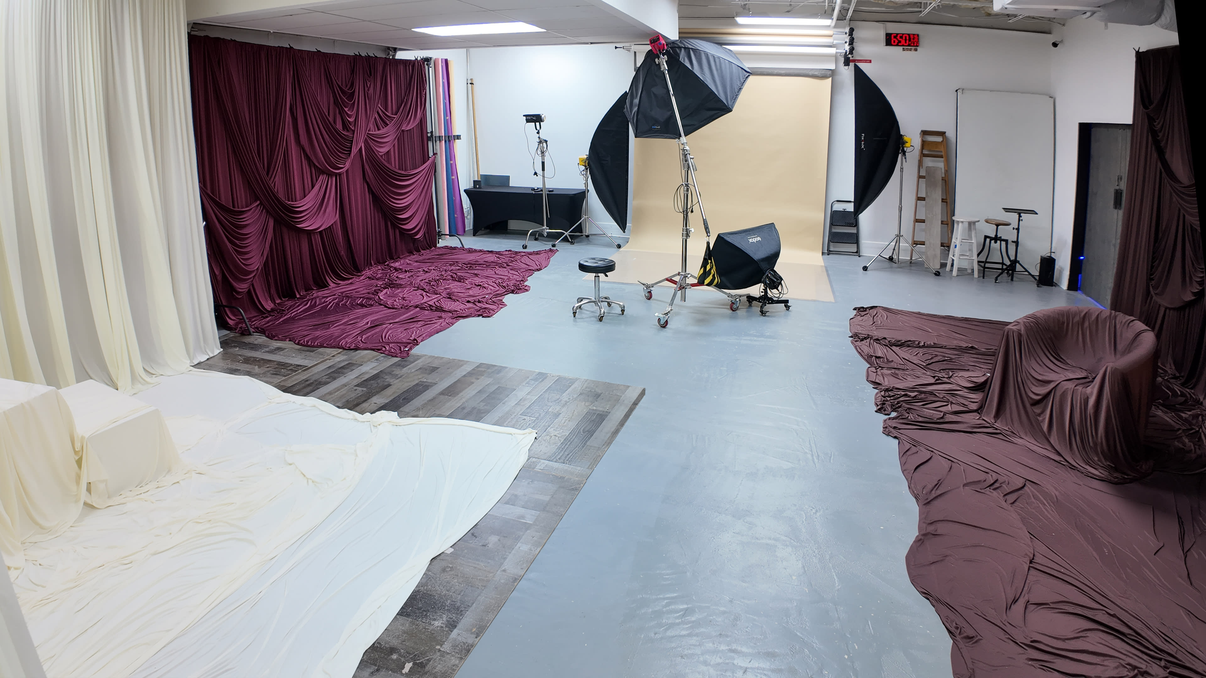 The image shows a photography studio with draped fabric in white and maroon, a gray floor, a backdrop setup, and various studio equipment including a light stand and a stool.