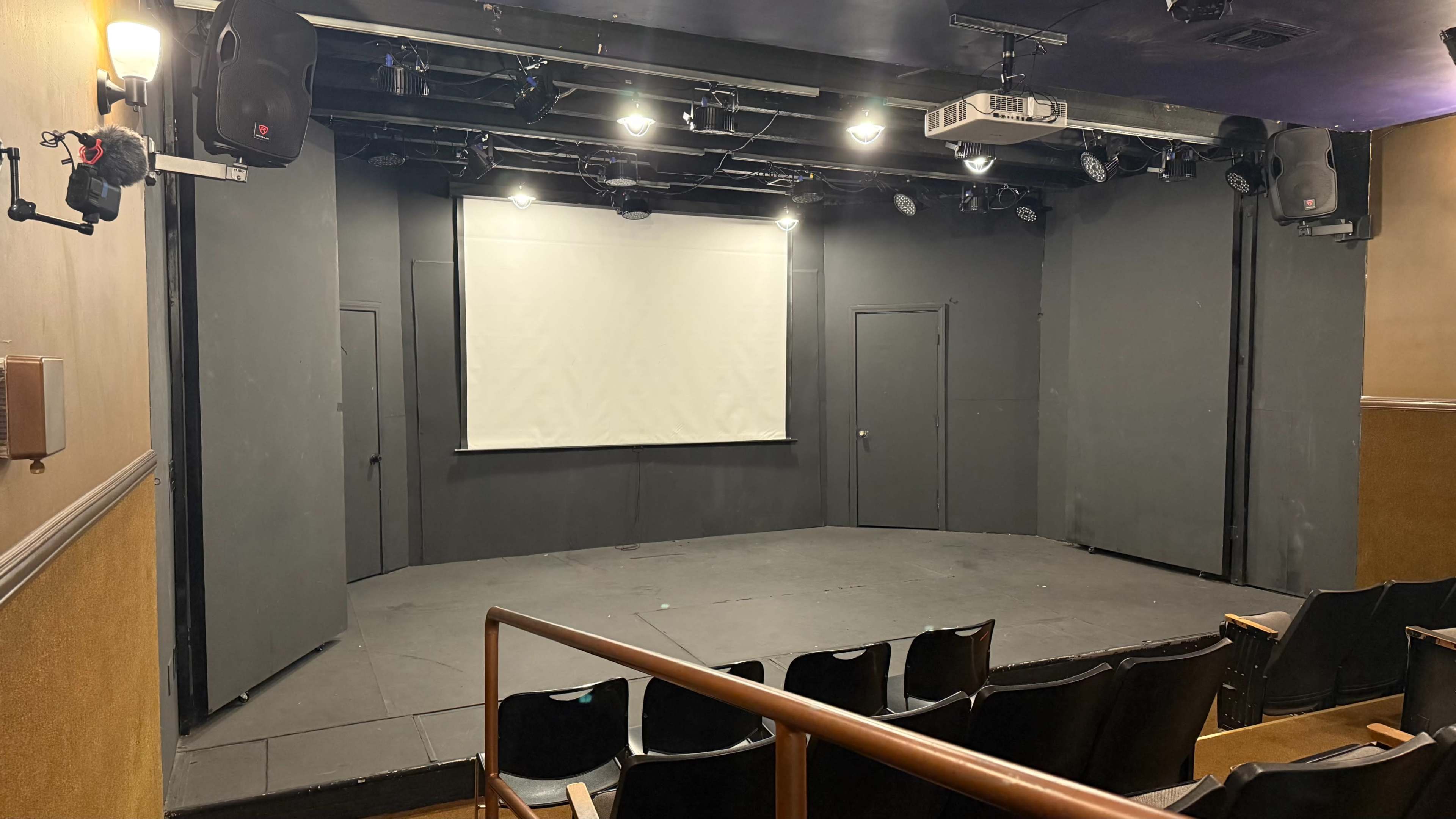 The image shows an empty theater stage with a projection screen and black walls, seating positioned in front.