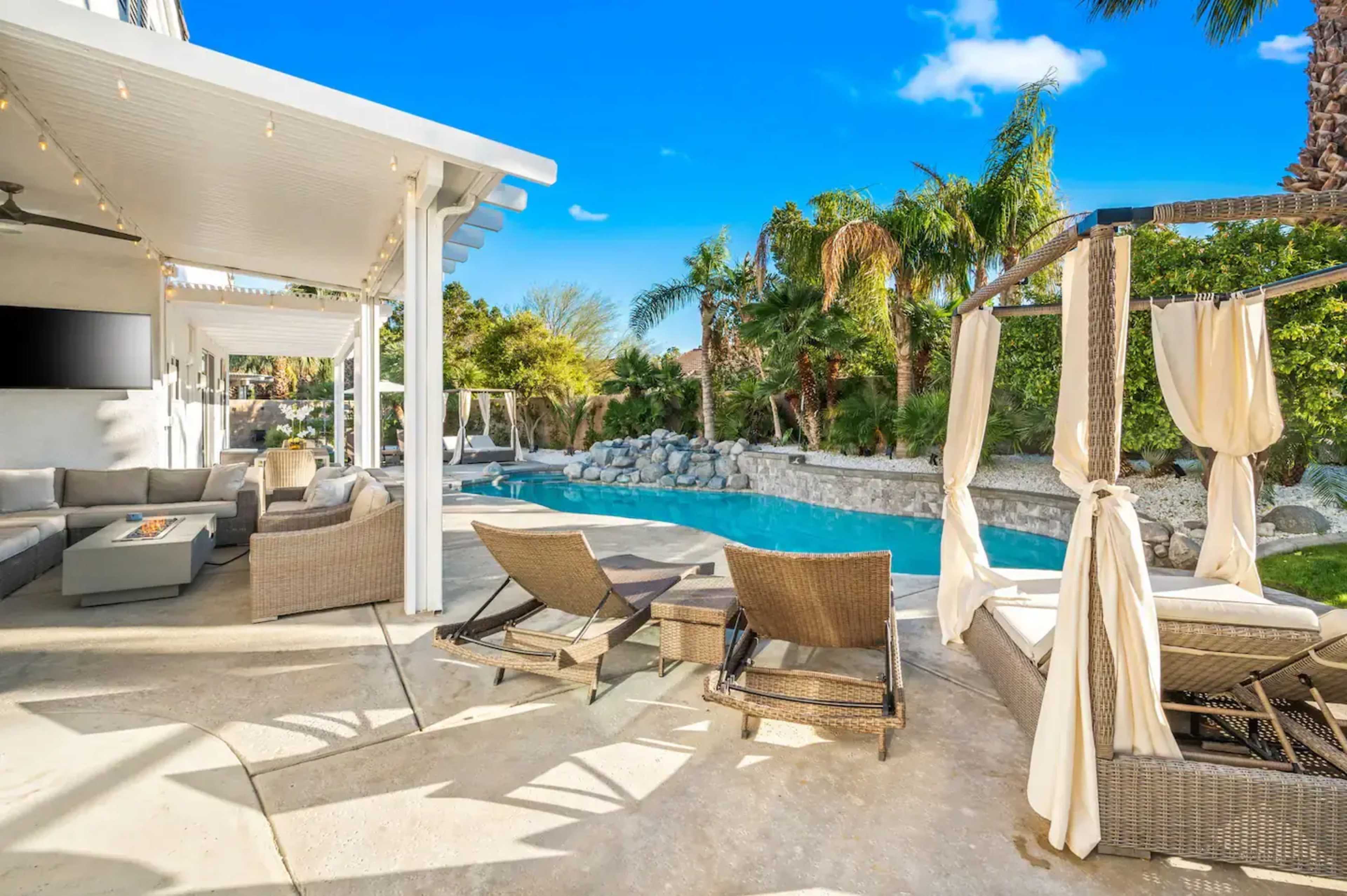 A backyard space with a swimming pool, lounge chairs, and a covered patio area surrounded by palm trees and landscaping.