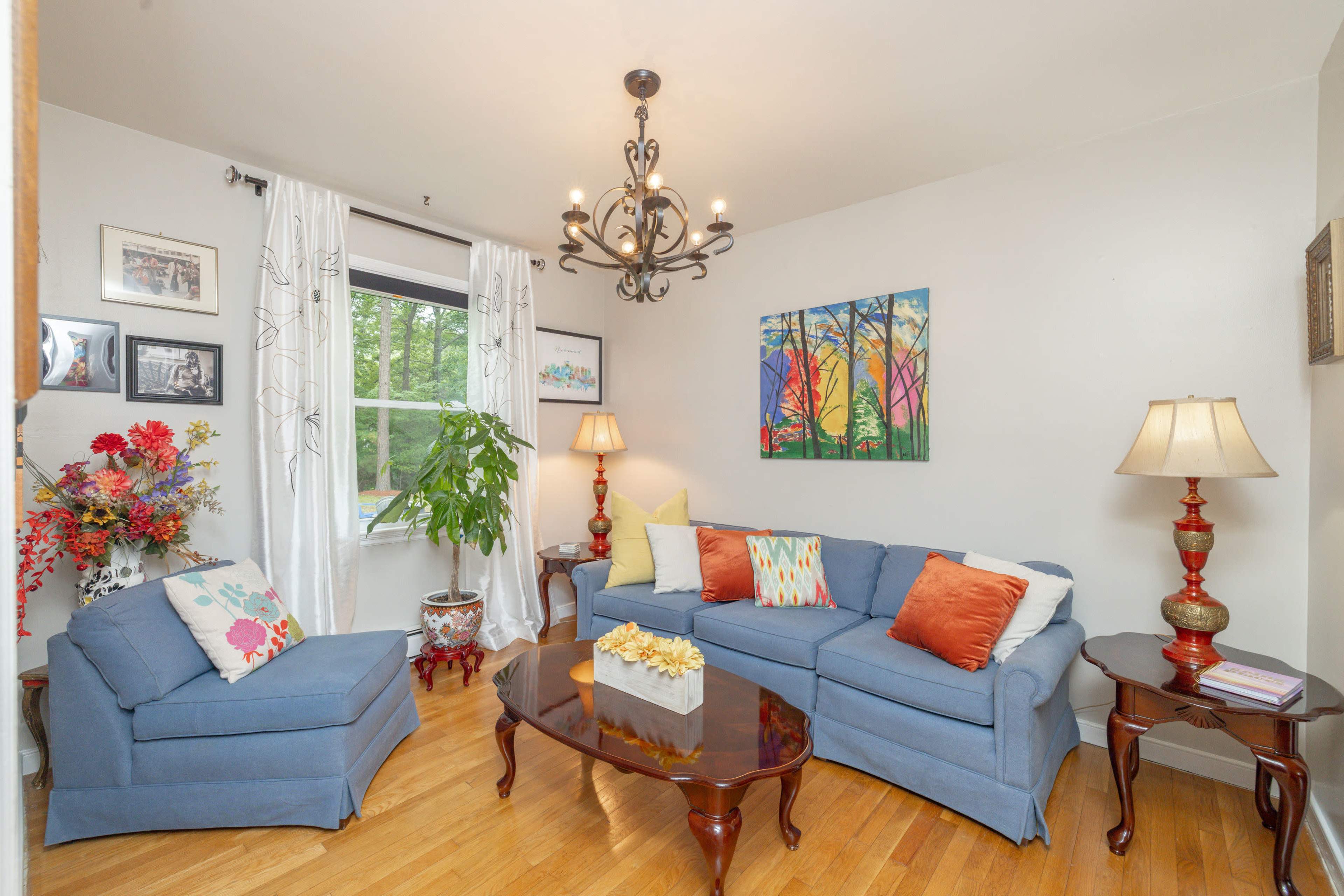 The living room features a blue sofa with colorful pillows, a wooden coffee table, two lamps, and a large window with sheer curtains.
