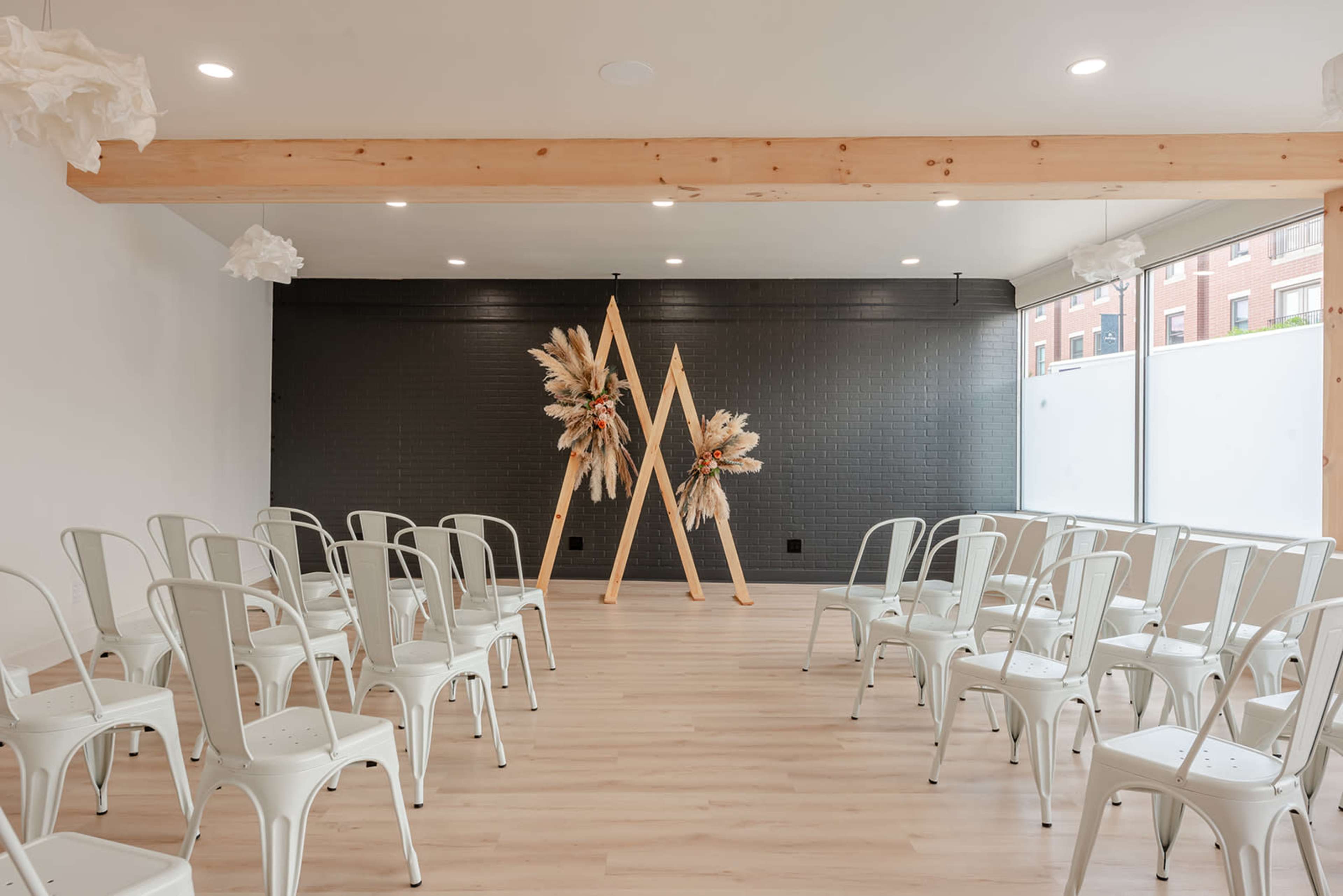 A modern event space features white metal chairs arranged in rows facing a decorative backdrop of wooden structures and dried floral arrangements.