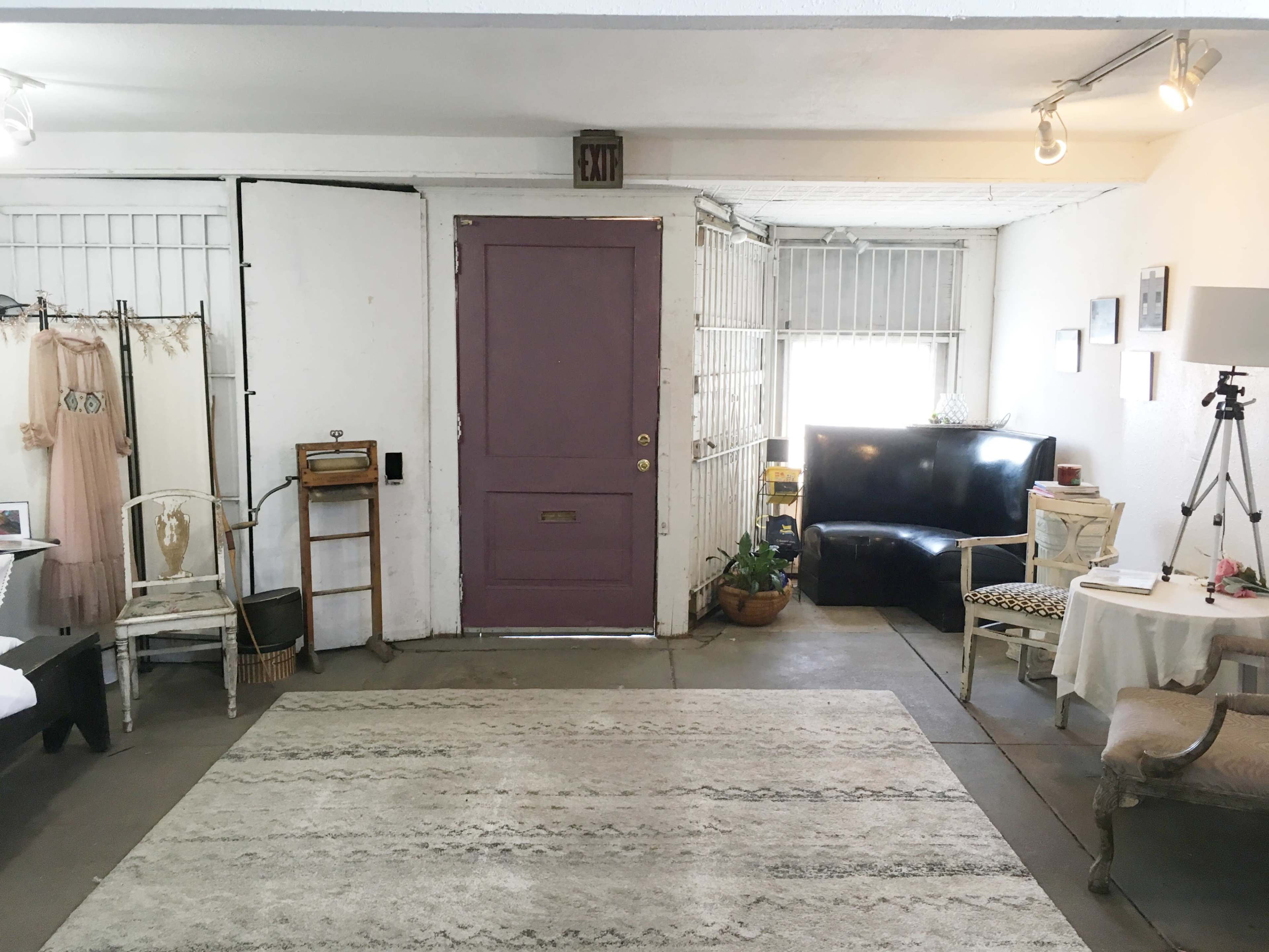 The image shows a room with a purple door, a black couch, a small table with a lamp, and various clothes hanging on the wall.