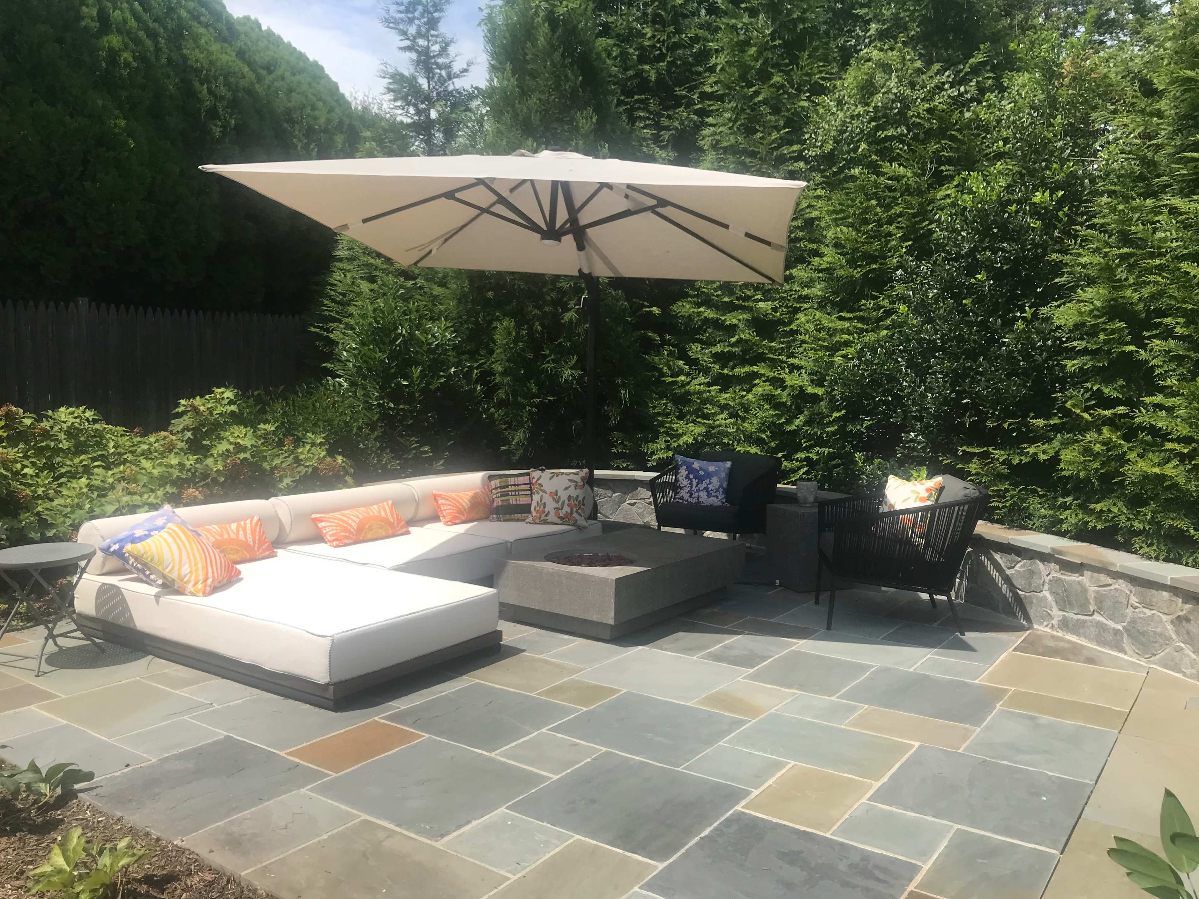 A patio area features a sectional sofa with colorful pillows, a central coffee table, and a large umbrella, surrounded by greenery and stone flooring.