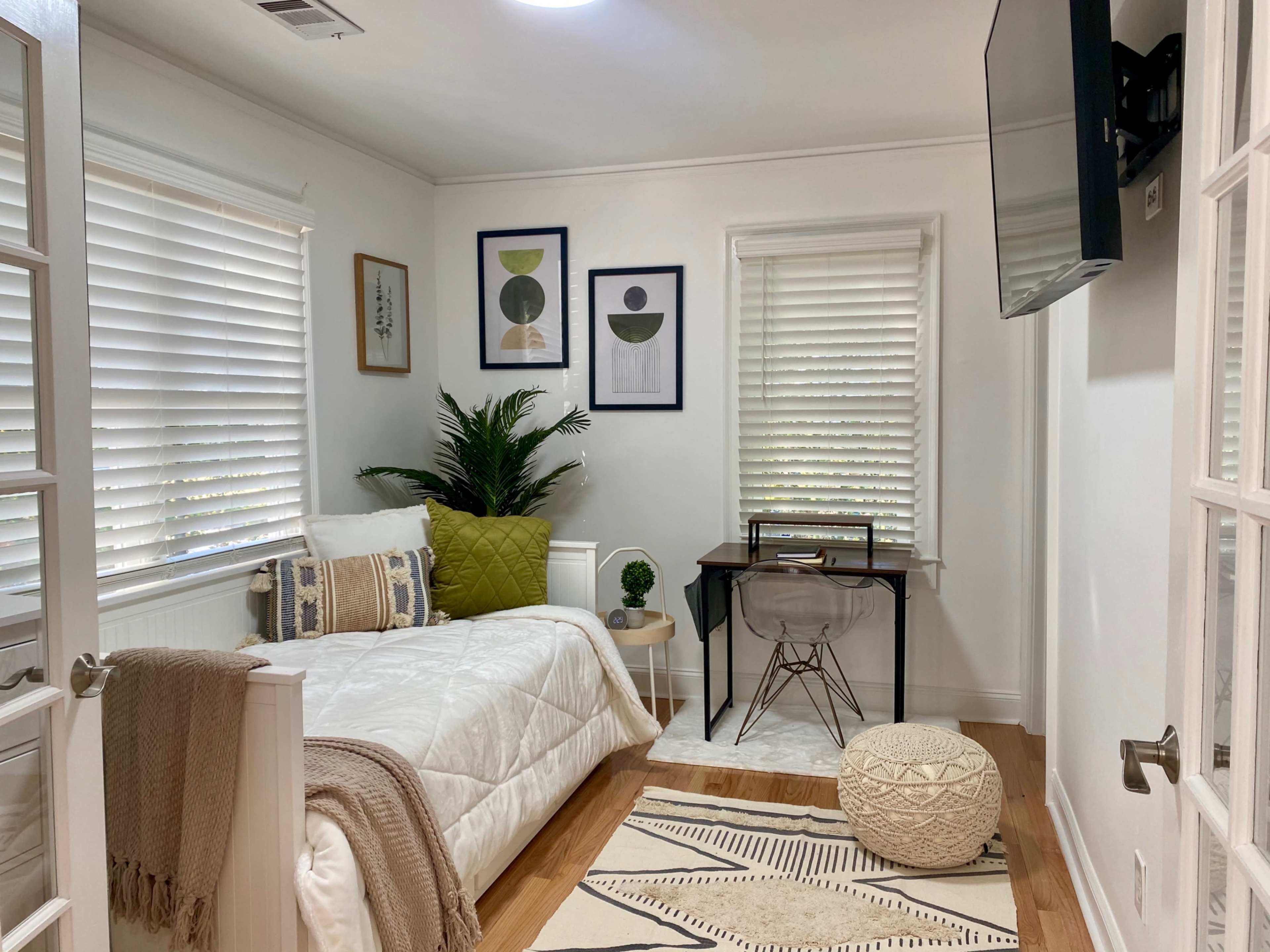 A small, well-decorated room featuring a bed, a desk with a chair, framed wall art, and a plant near a window.