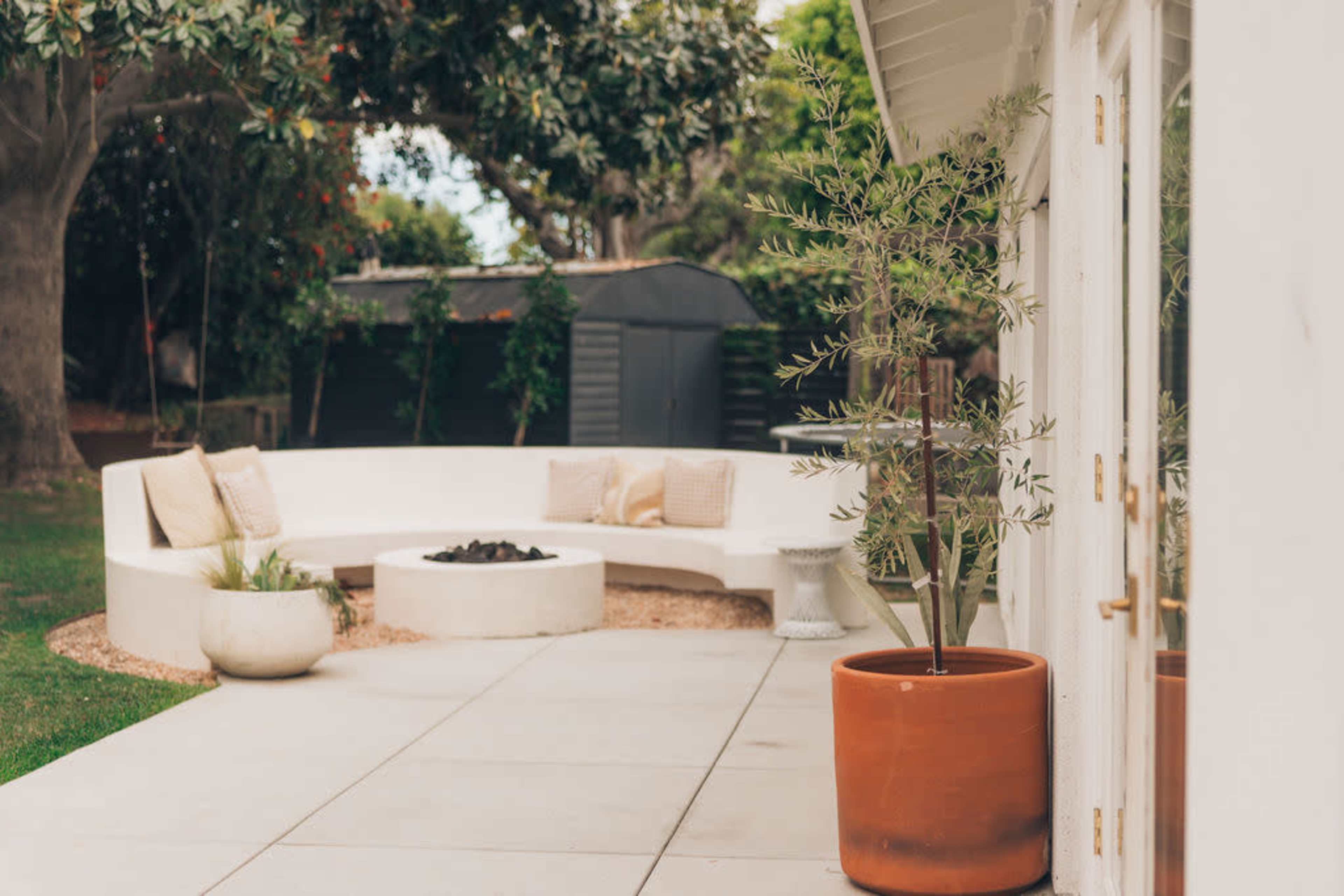 The image shows a modern outdoor patio with a curved sofa, a fire pit, and various potted plants.